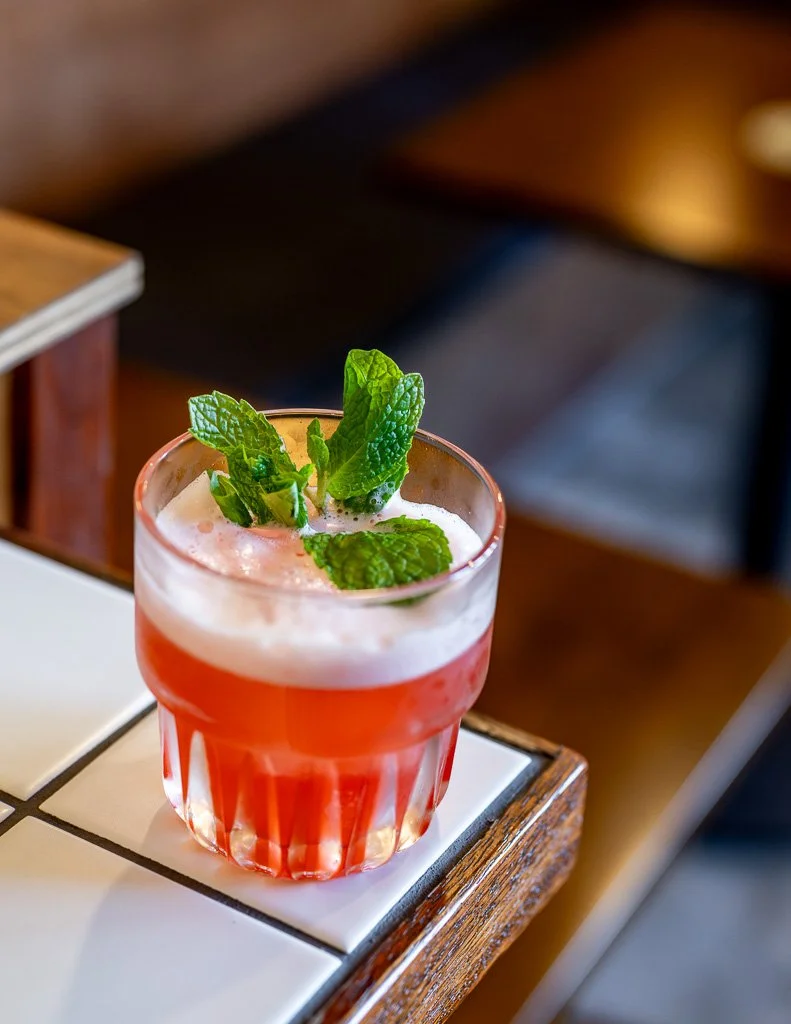 A glass of pink cocktail garnished with fresh mint leaves, placed on a tiled surface at a wooden table in a cozy setting.