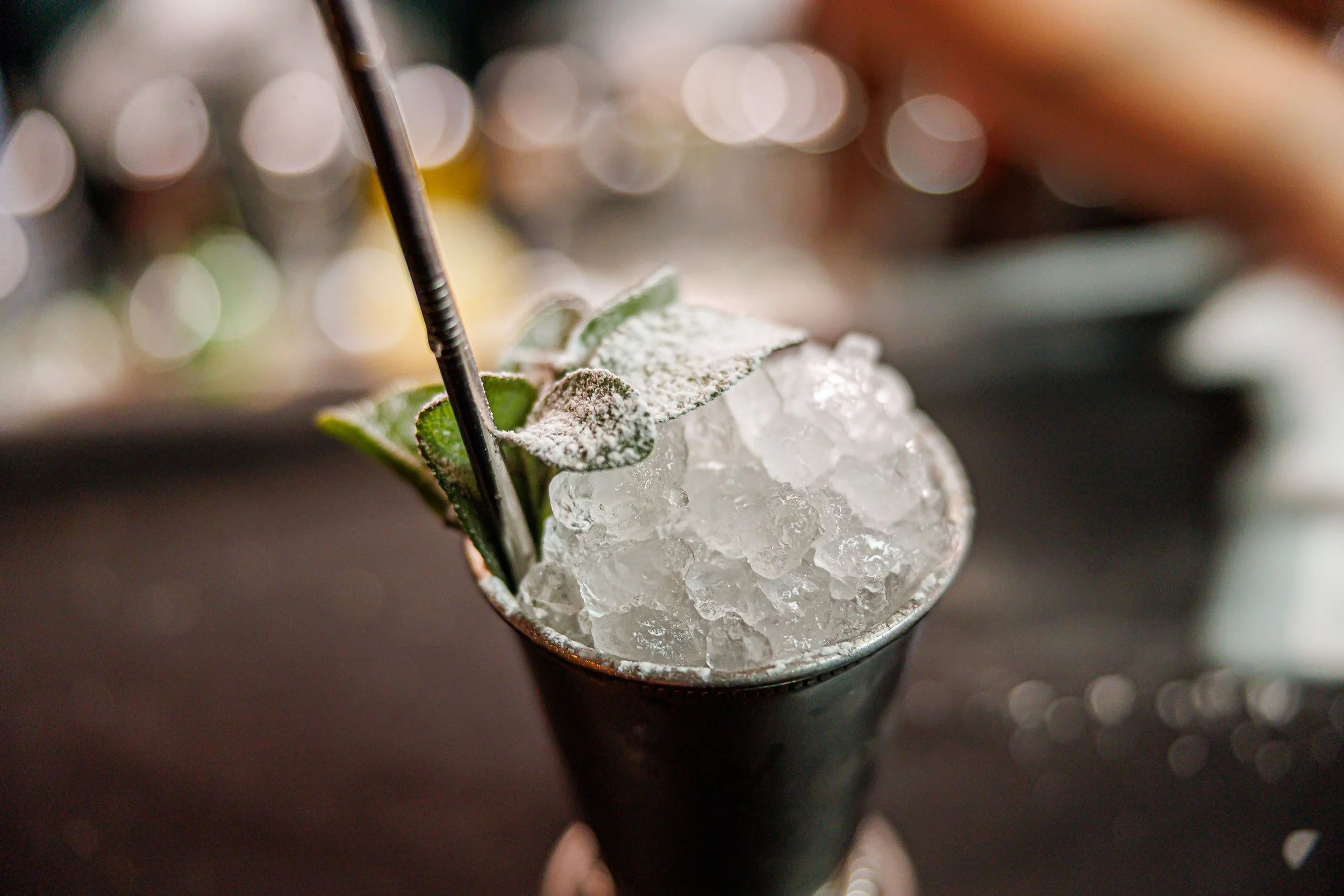 A cocktail in a metal cup filled with ice, garnished with a mint sprig dusted with powdered sugar and a black straw.
