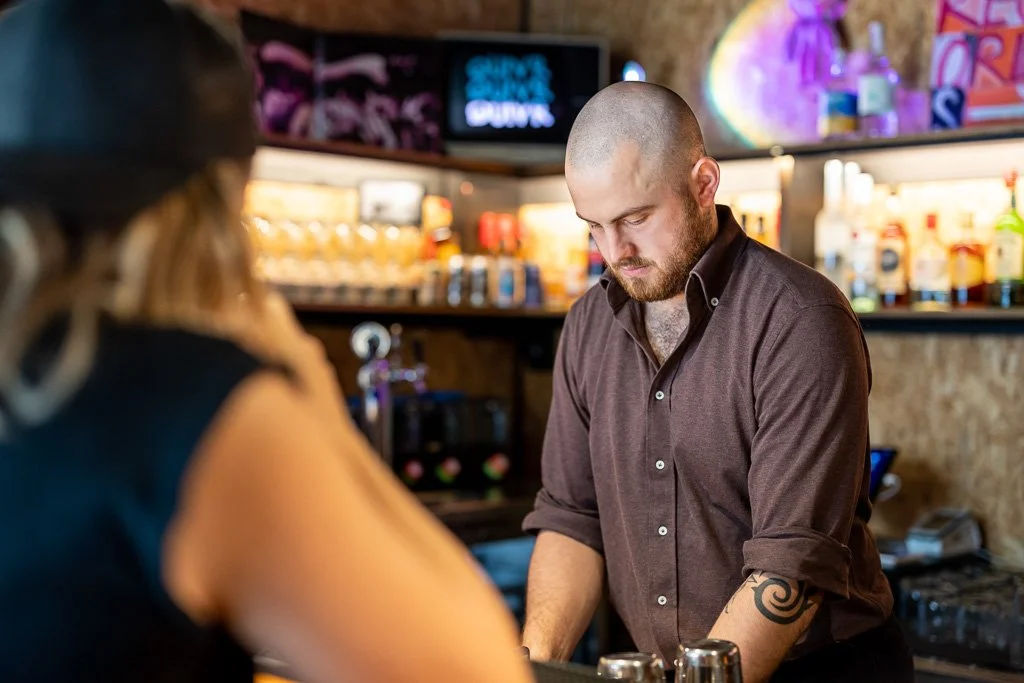 Bartender with a shaved head and tattooed arm serving drinks at a bar counter.