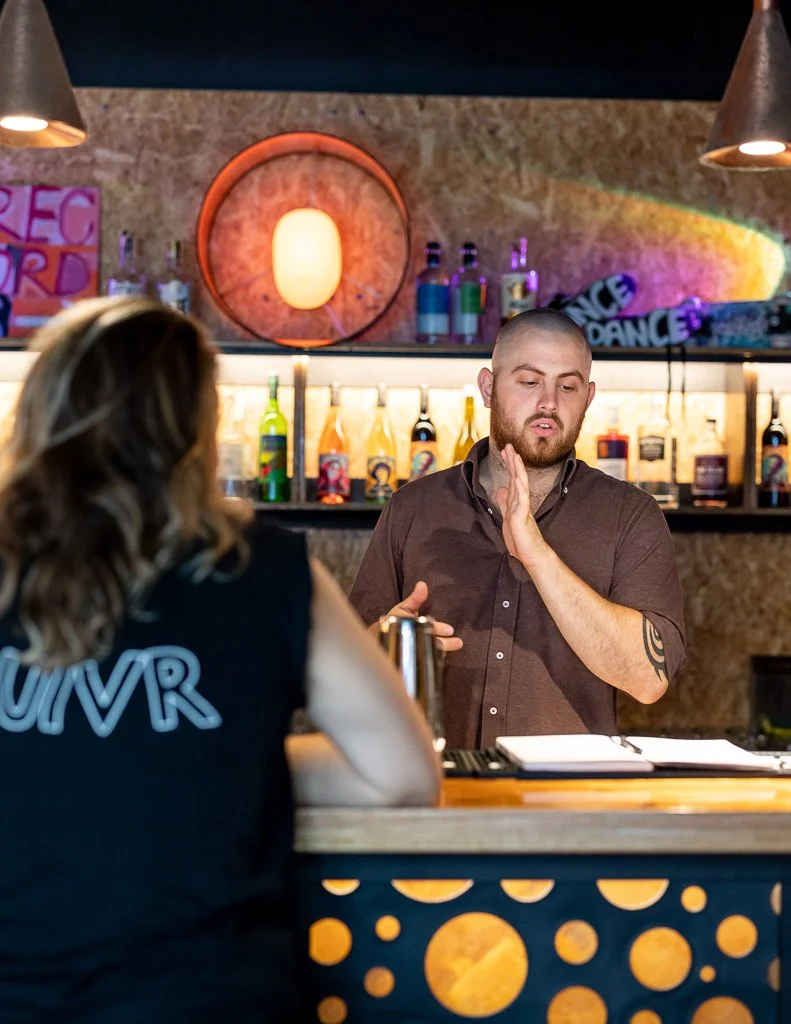 A man with a shaved head and beard, wearing a brown shirt, is behind a bar speaking to a woman with long wavy hair, wearing a black shirt with neon blue writing. The bar has liquor bottles on shelves, and the background features bar decor with colorf