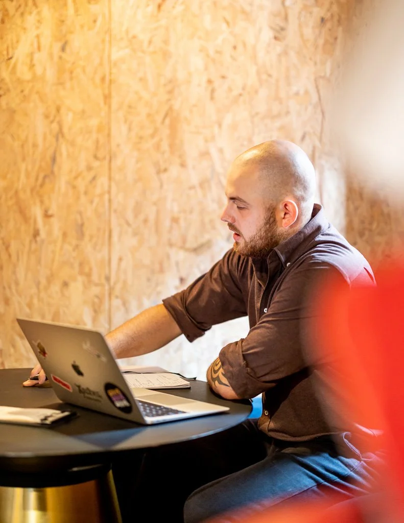 A man with a shaved head and beard working on a laptop at a table in a room with wood-paneled walls.