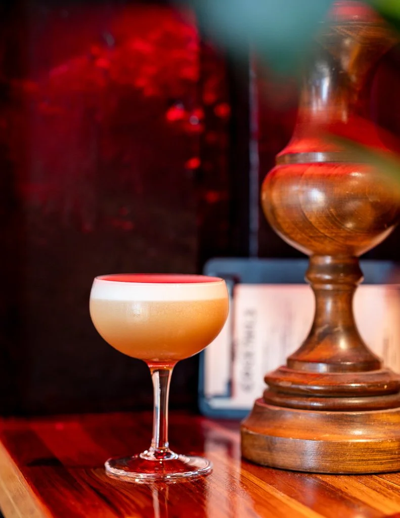A cocktail in a coupe glass on a wooden bar countertop, with a large wooden decorative object in the background.