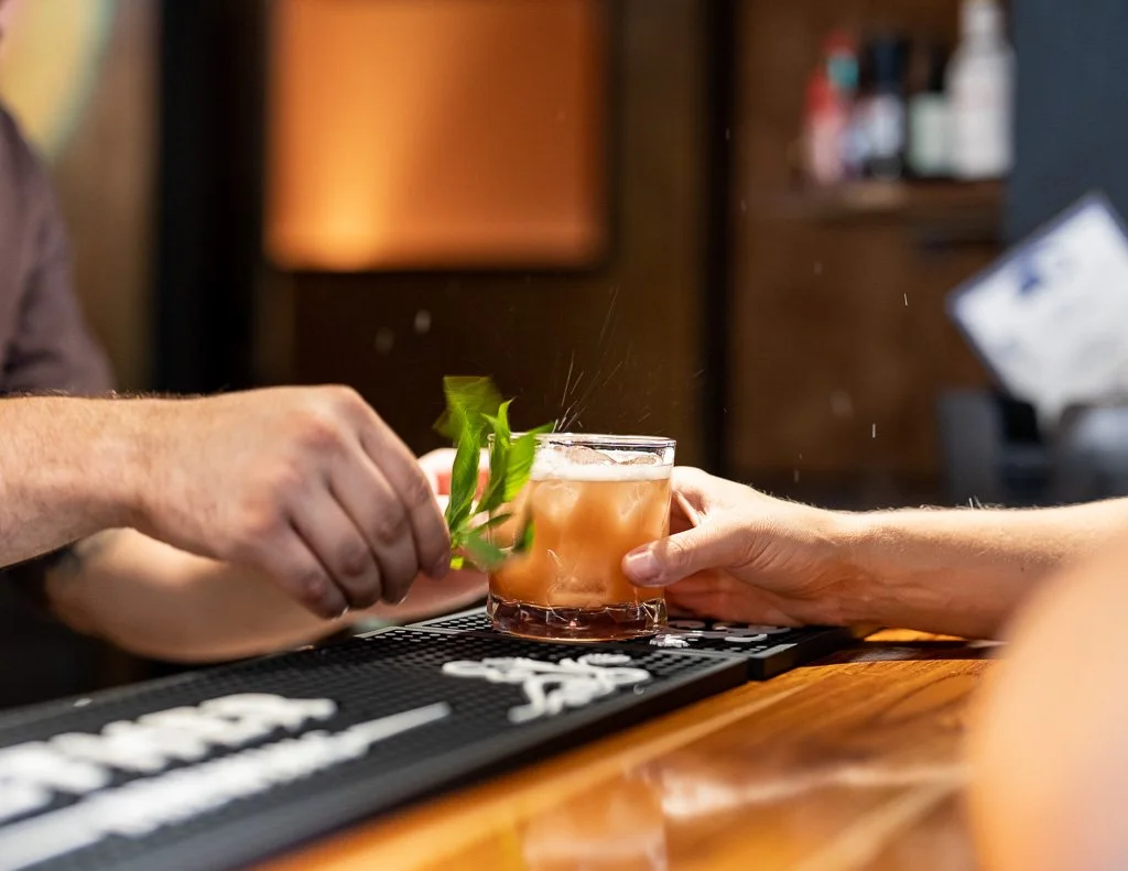 A bartender is garnishing a cocktail with a sprig of mint, while a person in front holds the glass on a bar counter.