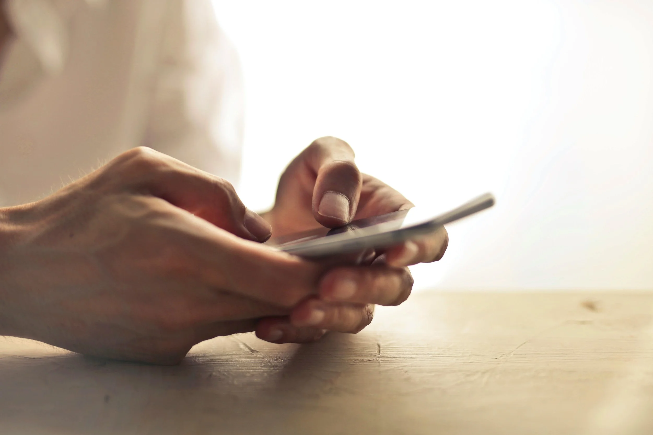 Close-up of hands holding and using a smartphone on a wooden surface with a blurred background.