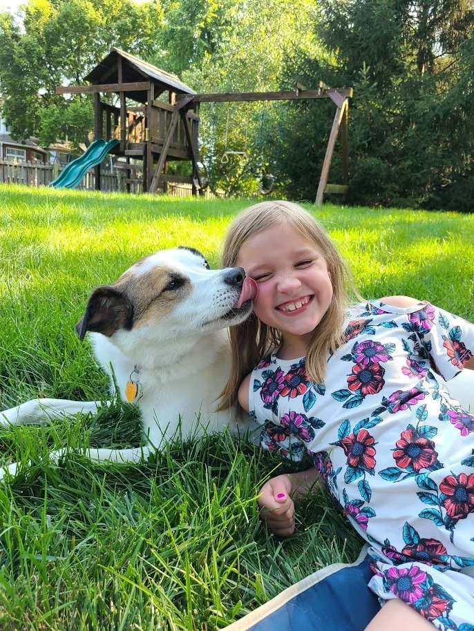 A young girl lying on green grass smiling as a dog licks her face in a backyard with a wooden playset and swing set in the background.