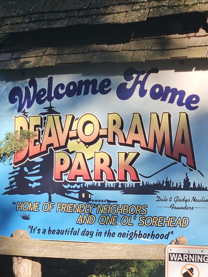 Colorful community sign for Deaver-Aroma Park welcoming visitors, featuring trees, a lake, and a house silhouette, with text indicating it as a friendly neighborhood and the home of 'One Ol' Sorehead'.