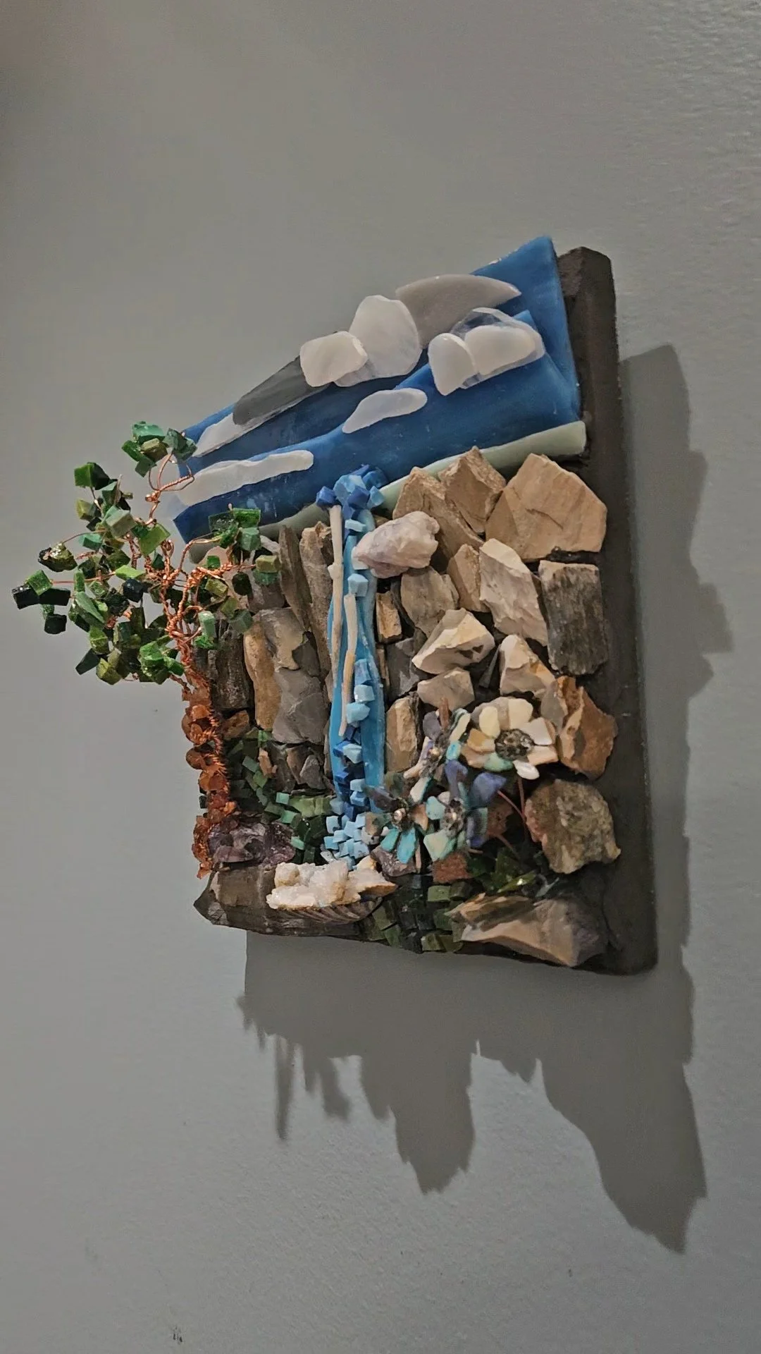 Mixed media art piece with a miniature waterfall, rocks, a small tree, and seashells on a wooden base.