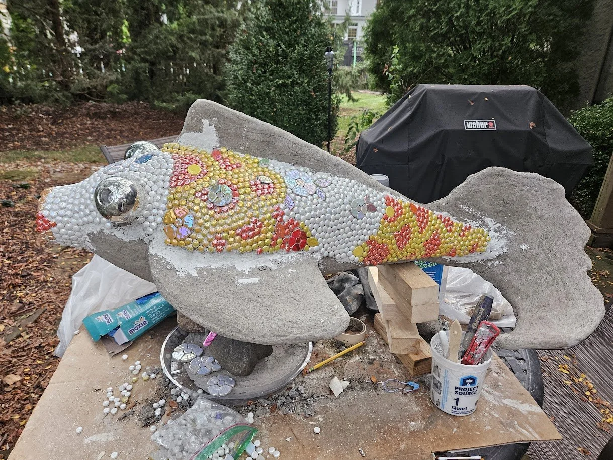 A concrete fish sculpture in progress decorated with colorful mosaic tiles and beads, situated outdoors on a workbench with tools and supplies around it.