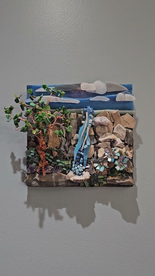 Mixed-media art piece depicting a landscape with a cloudy sky, mountain, and waterfall, featuring natural stones, painted clay, and small decorative elements.