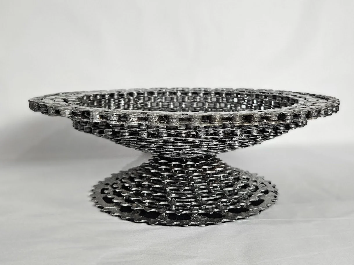 A metallic chainmail bowl composed of interconnected metal rings, arranged in a circular and layered pattern, with a flat base and a wide open top, set against a plain white background.