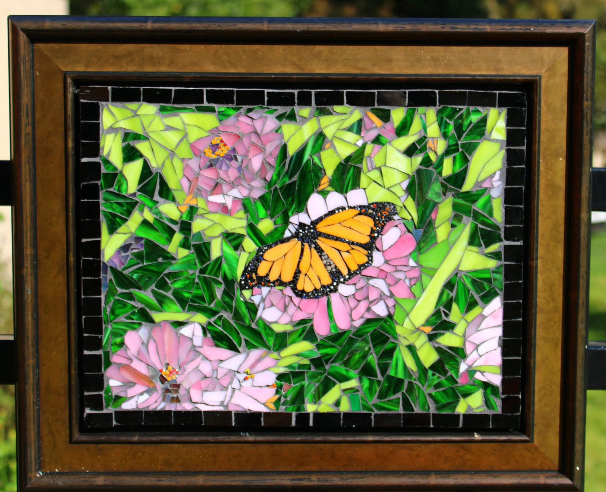 A framed mosaic artwork featuring pink flowers and an orange butterfly with black and white accents, set against a background of green leaves and grass.