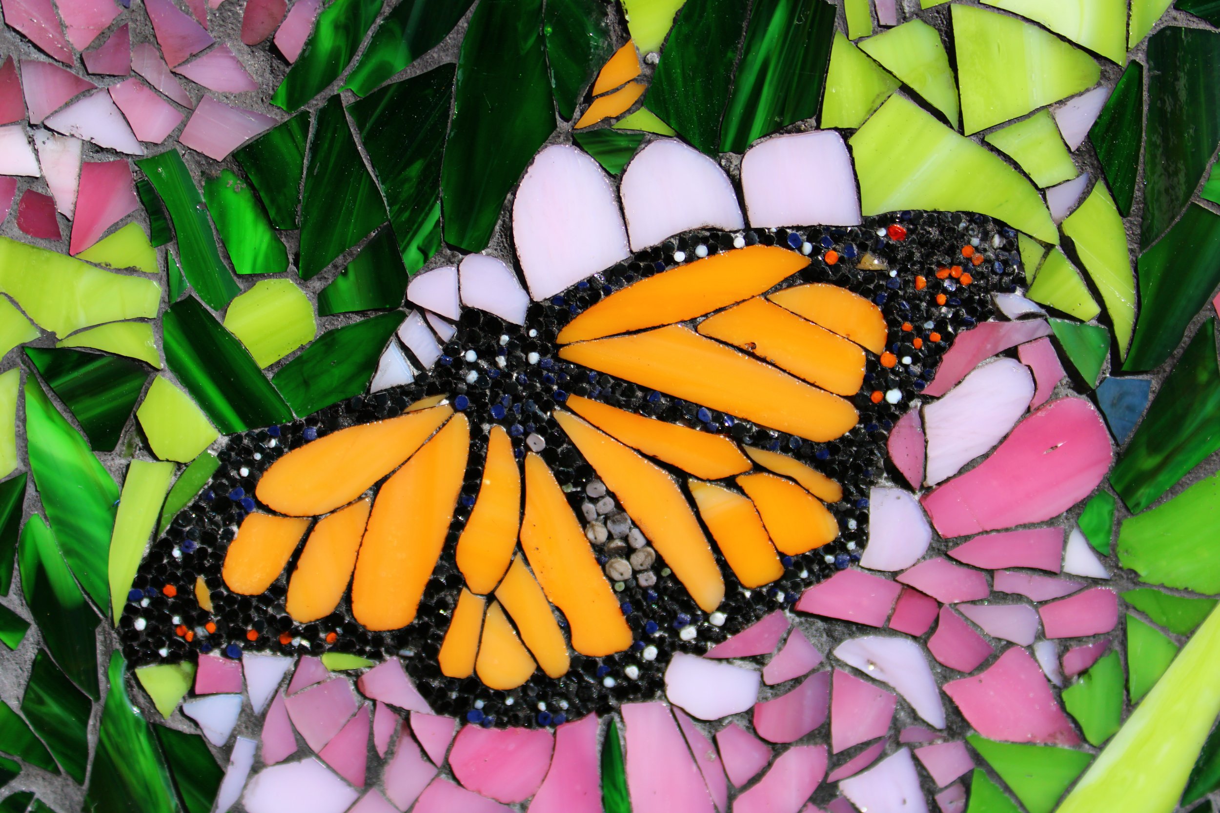 Close-up of a colorful butterfly mosaic made of small pieces of glass and tile, with orange wings and black details, situated among green, pink, and white tiles.