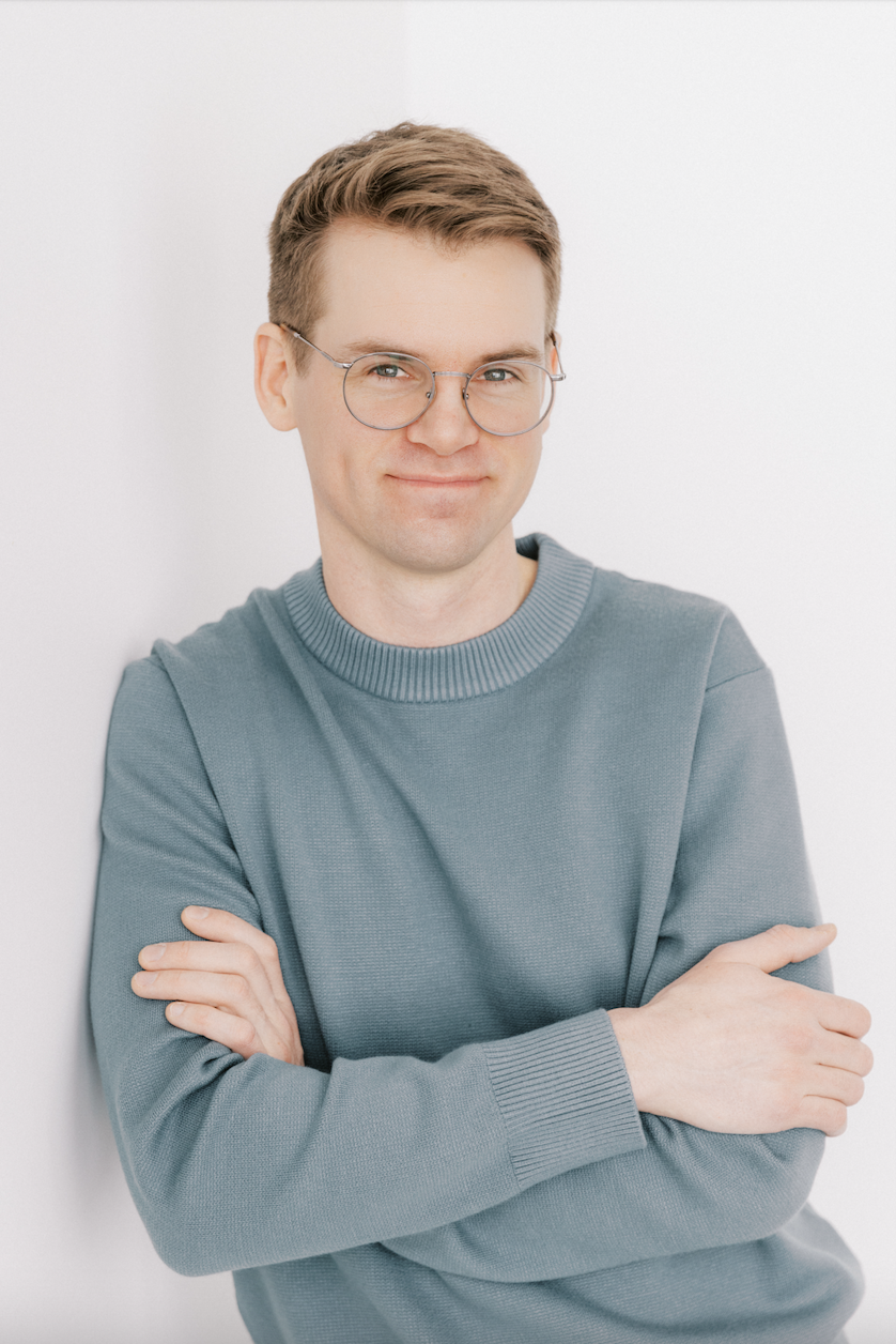 A young man with short light brown hair and glasses, wearing a gray sweatshirt, standing with arms crossed against a plain white background.