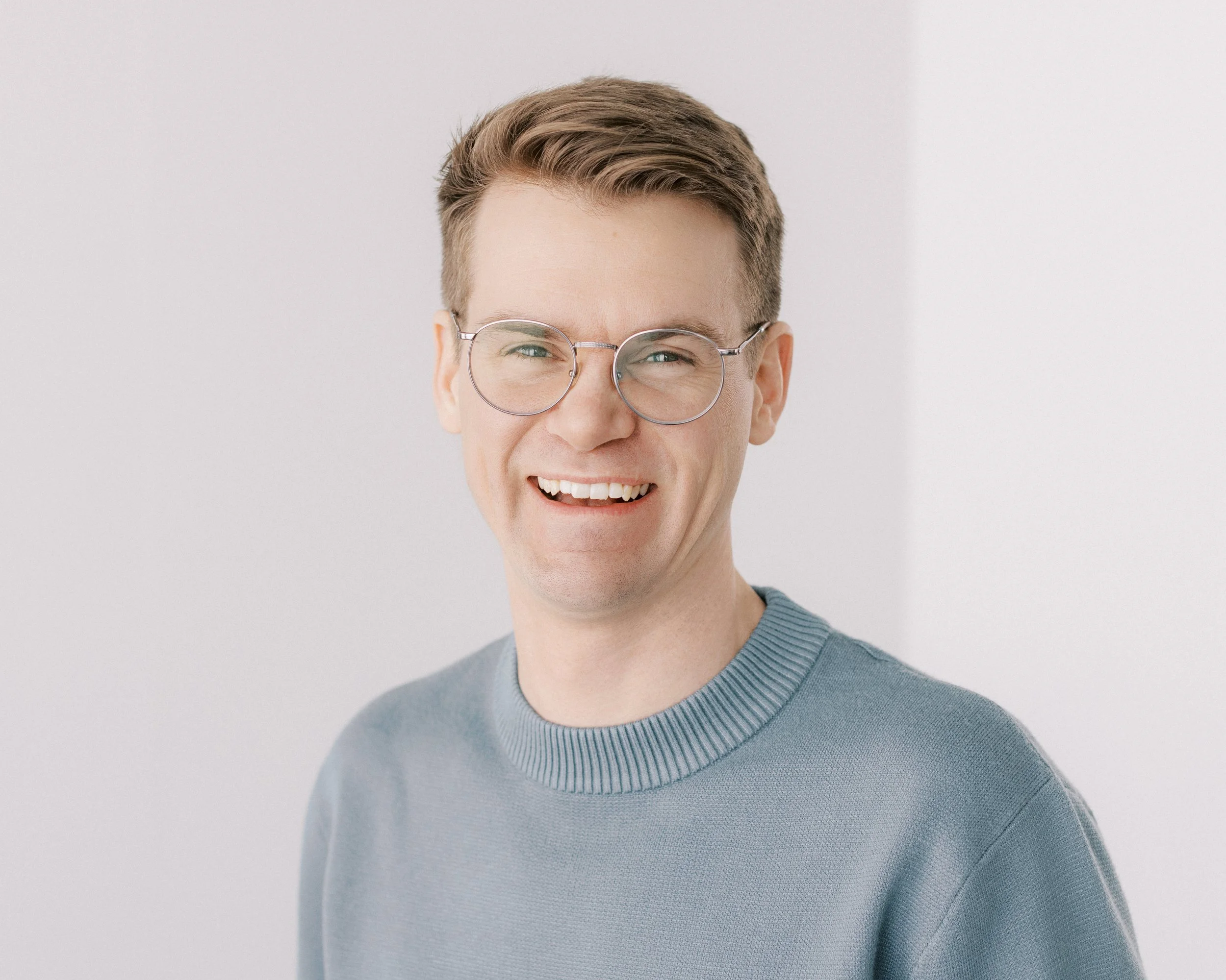 A young man with glasses, wearing a light blue sweater, smiling against a plain light background.