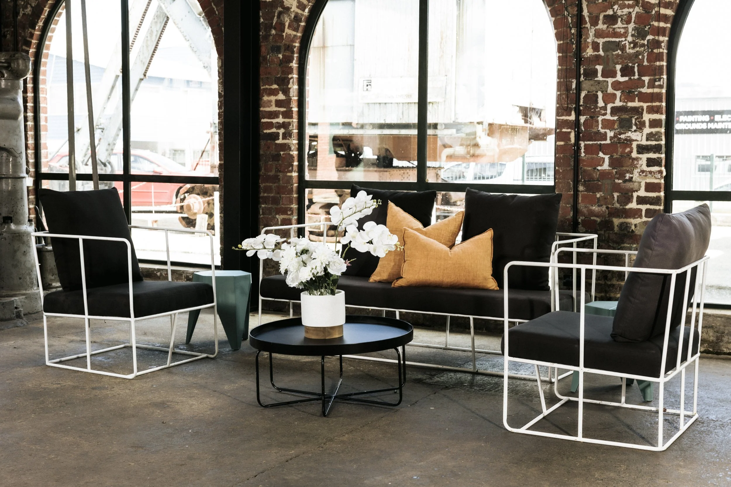 Interior of a modern industrial-style lounge area with black and white furniture, a black coffee table with a vase of white flowers, and brick walls with large arched windows.