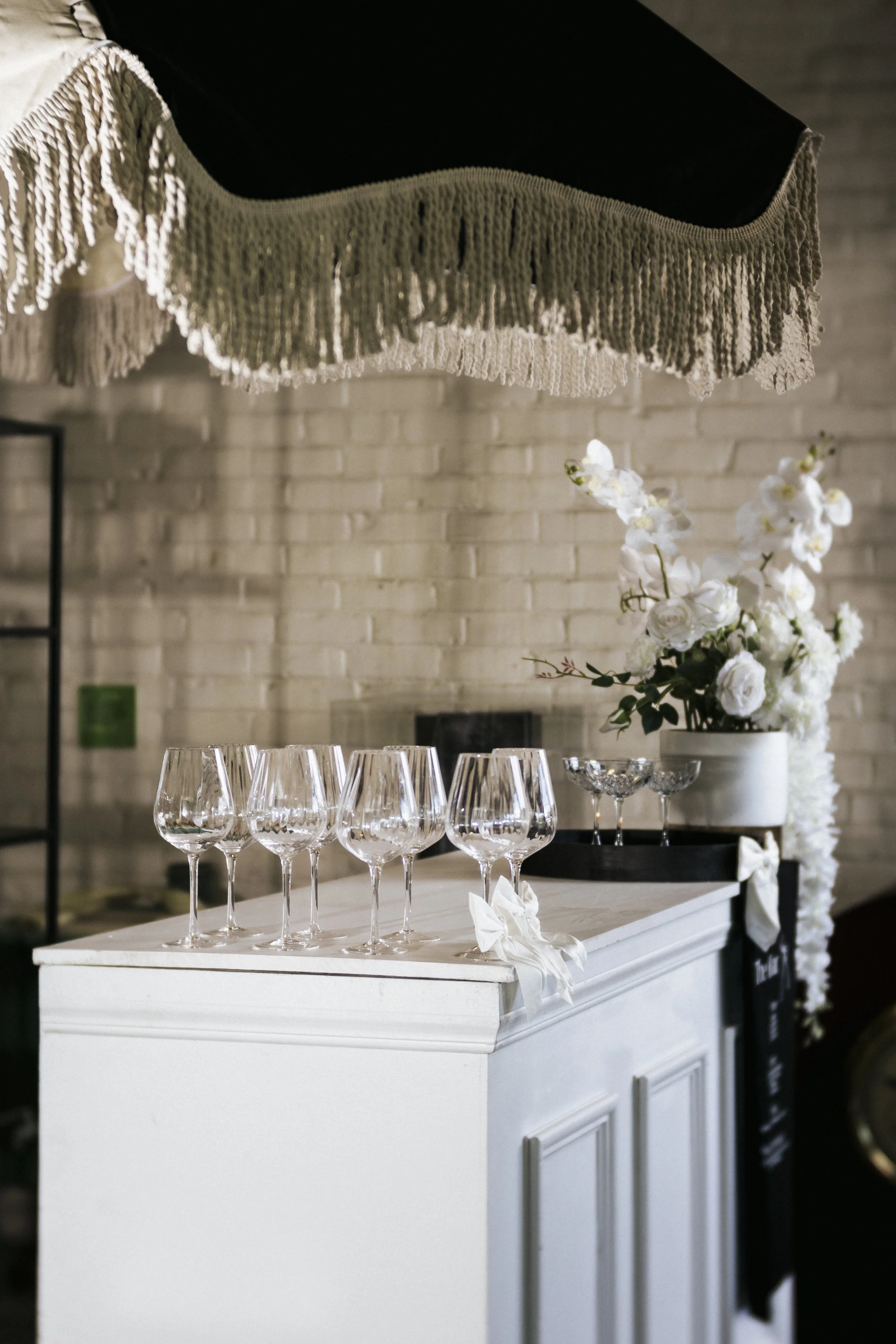 Empty wine glasses on a white bar counter with a white flower arrangement in the background, under a fringed lampshade.