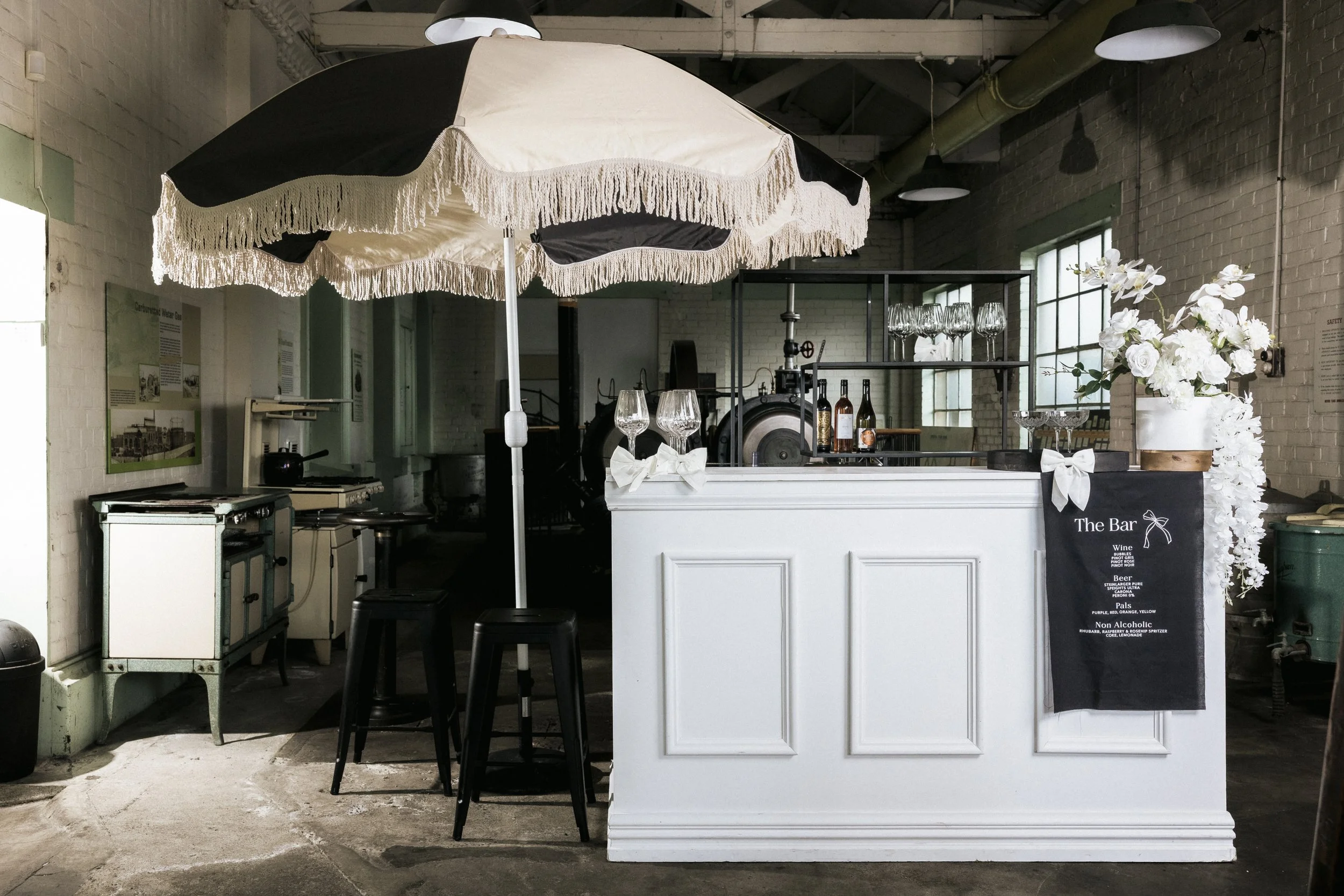Indoor bar setup with a large black-and-white umbrella, white bar counter decorated with white bows, wine glasses, and a vase of white flowers, with a chalkboard menu listing wine, beer, spirits, and non-alcoholic drinks, in a rustic industrial space with brick walls and large windows.
