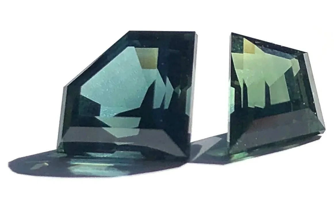Two large, dark green, Australian sapphire gemstones with sharp edges and geometric cuts, casting subtle shadows on a white surface.