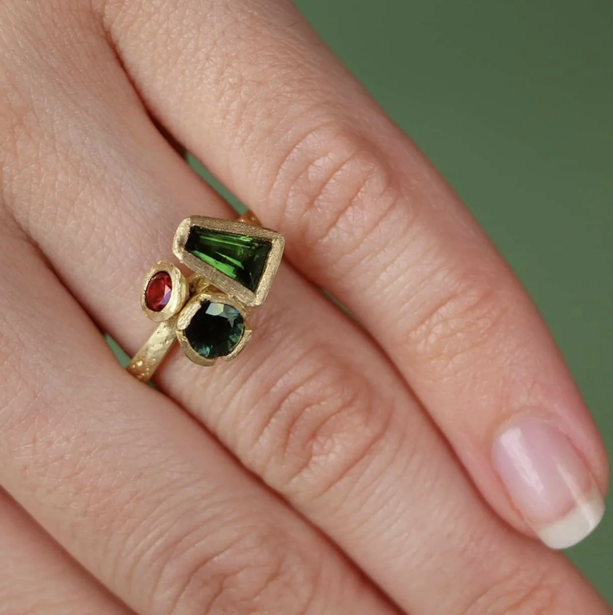 Close-up of a person's finger wearing a gold ring with three colorful gemstones: a green, a red, and a dark green or black stone.