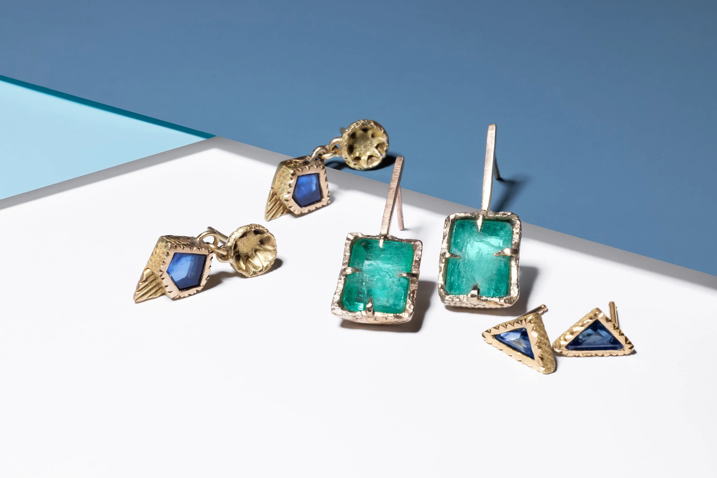 Collection of jewelry including earrings with blue gemstones and green gemstones, placed on a white surface with a background of blue and teal colors.