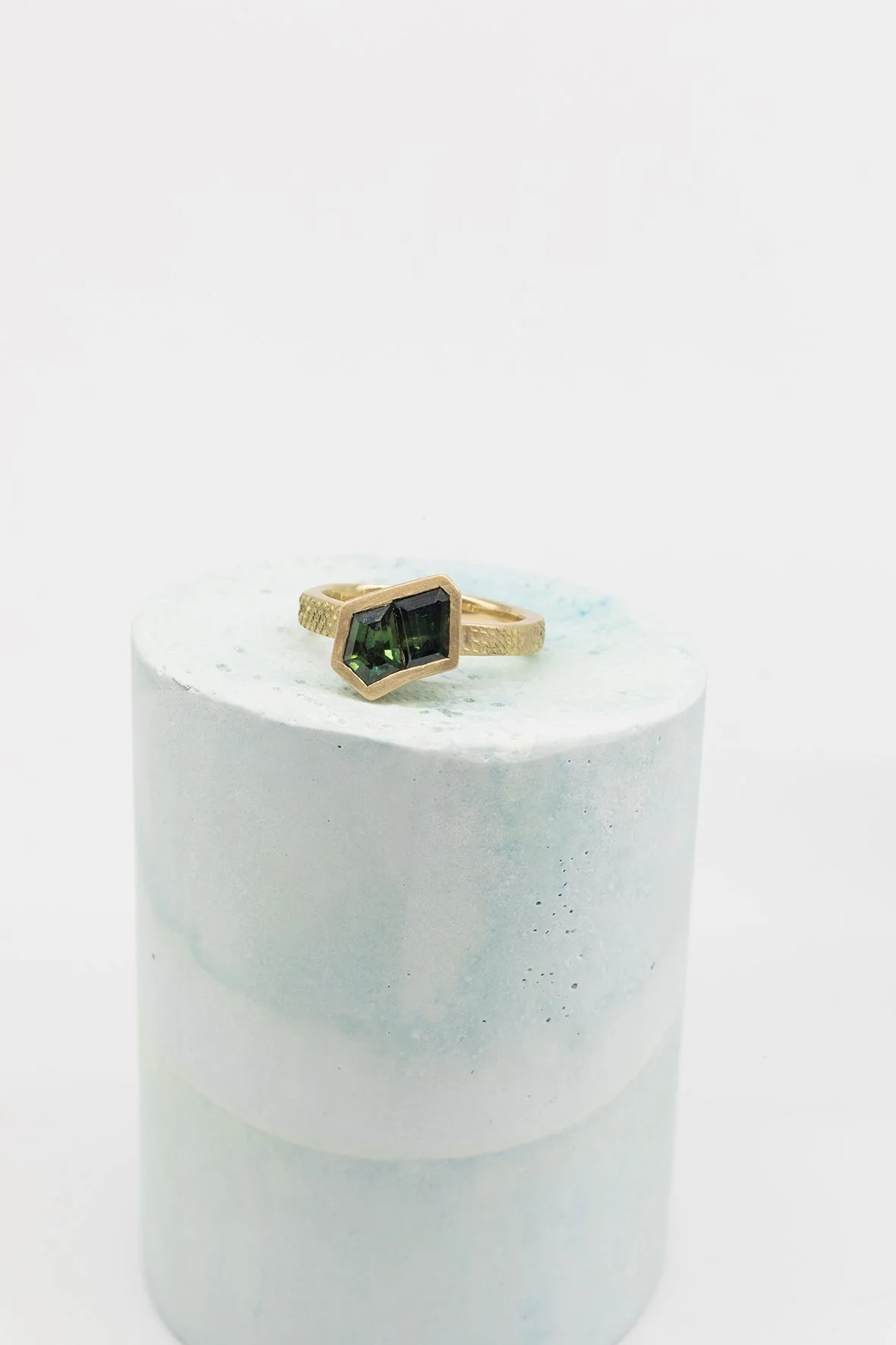 Gold ring with two dark green gemstones placed on a white cylindrical platform.