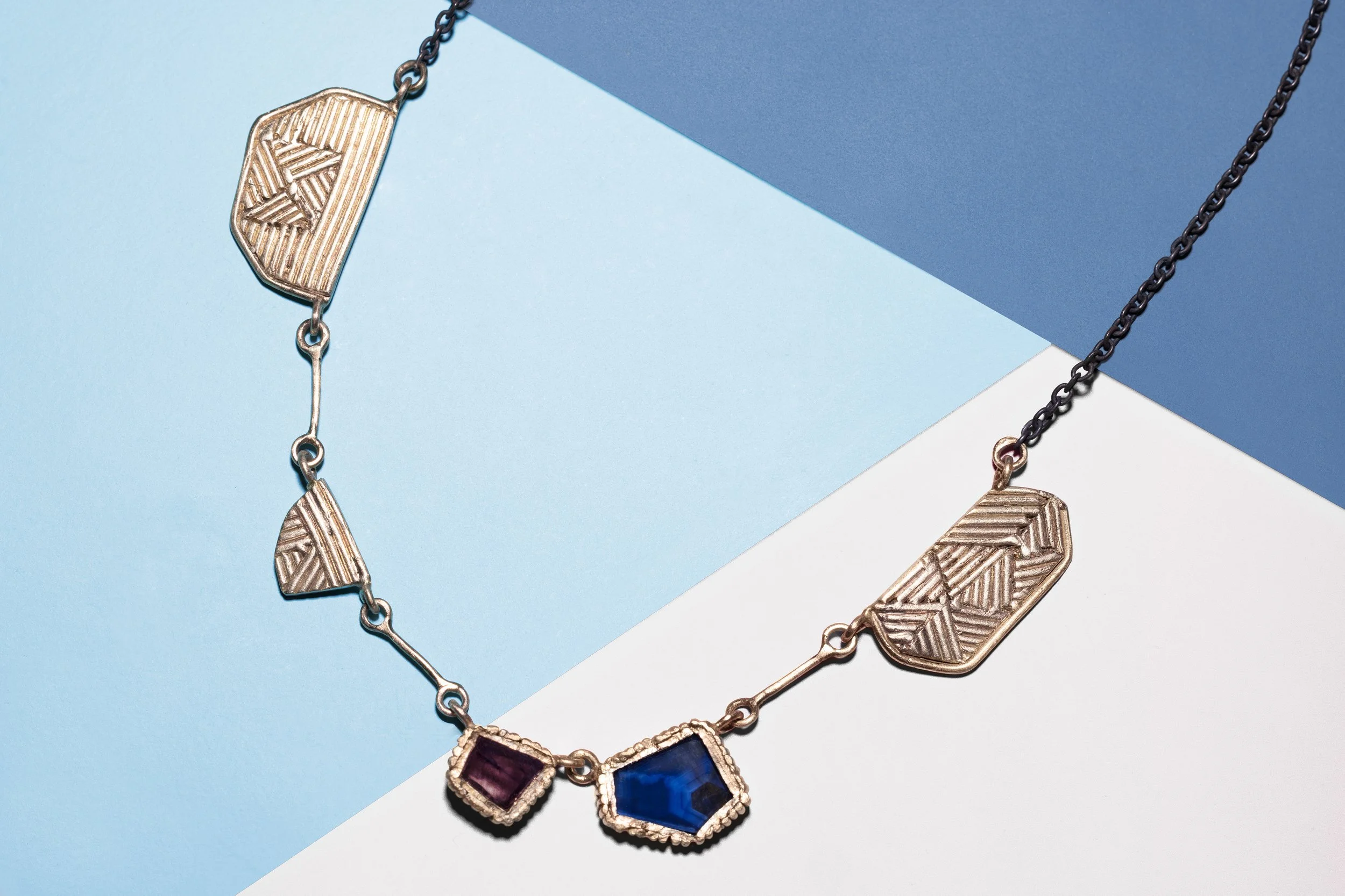 A necklace with geometric gold elements and two gemstone pendants, one purple and one blue, on a background of light blue, dark blue, and white paper.