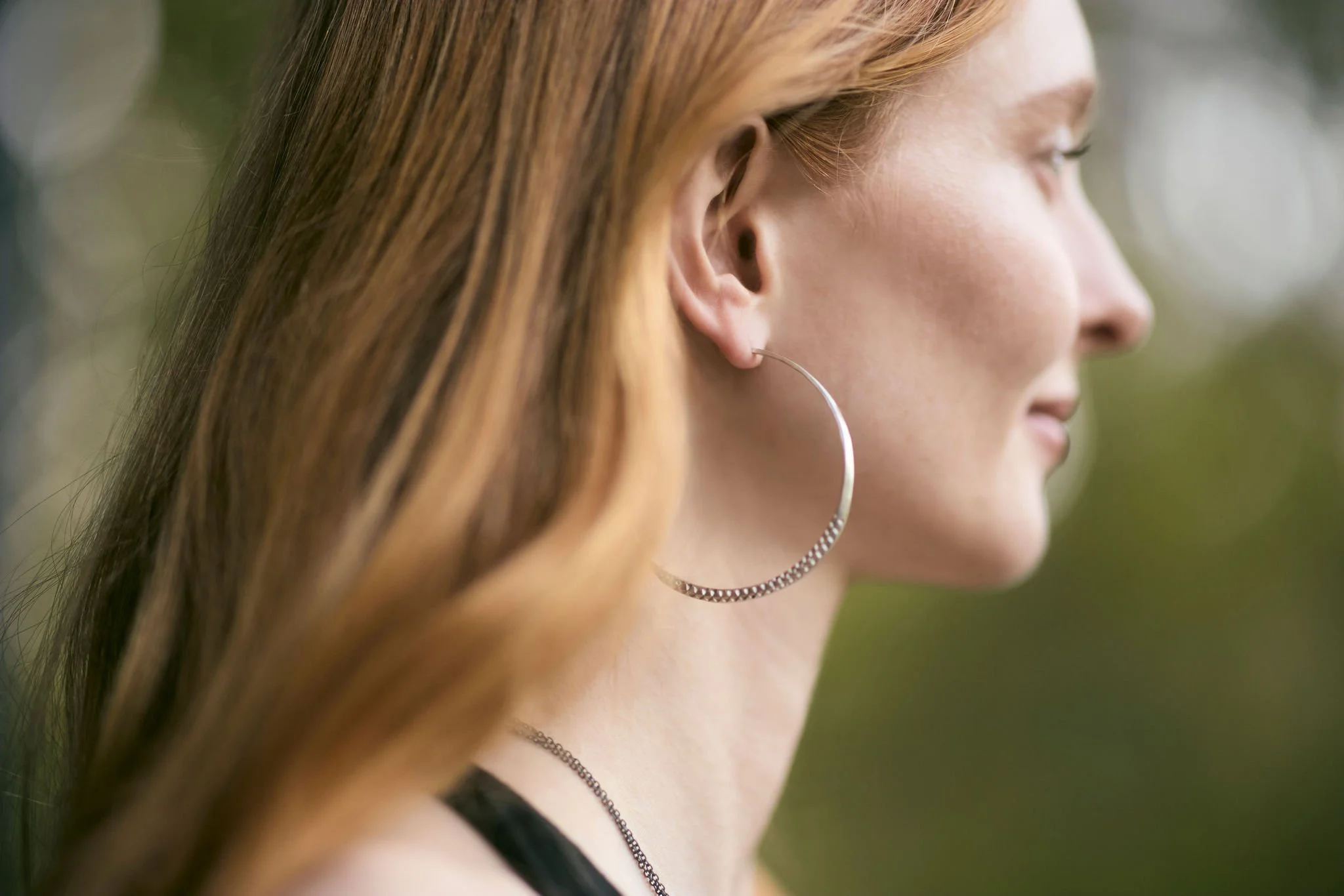 Close-up of a young woman with long reddish-brown hair, wearing a large hoop earring and a black top, outdoors with a blurred natural background.