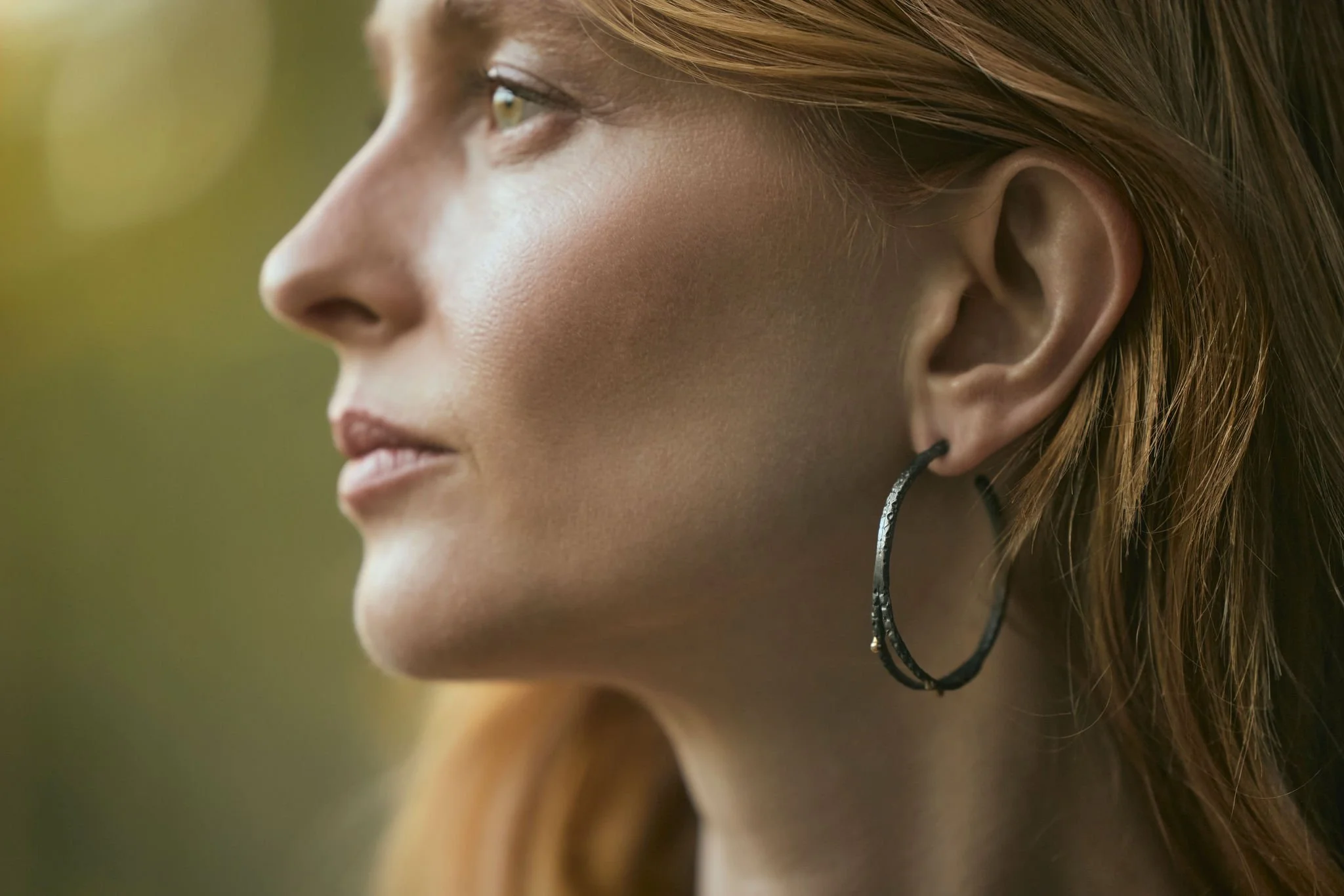 Close-up of a woman's face, focusing on her profile, with a large hoop earring, light makeup, and reddish hair, outdoors with a blurred natural background.