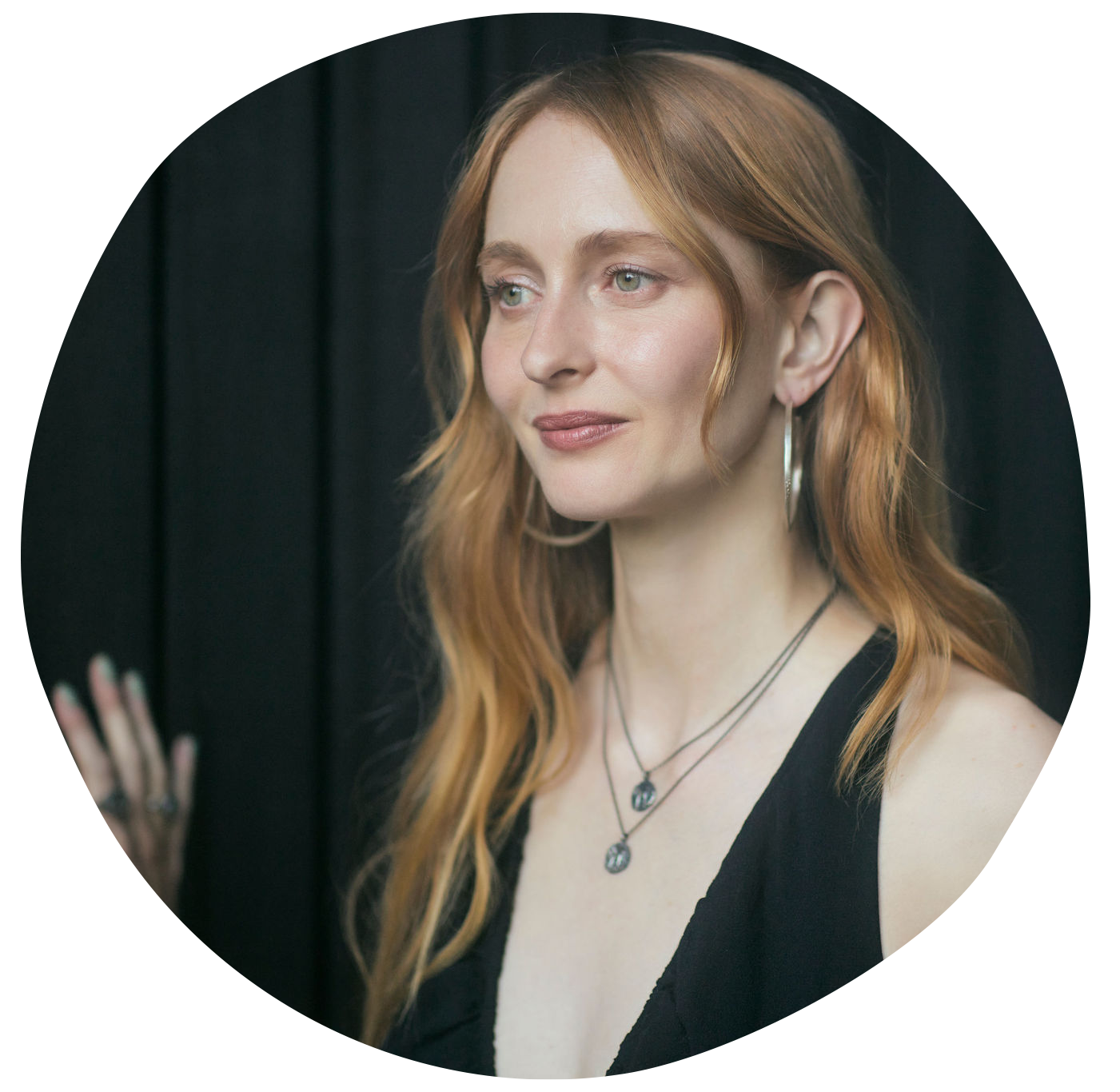 A woman with long, wavy red hair wearing hoop earrings, layered necklaces with pendant charms, and a black sleeveless top standing against a dark background.