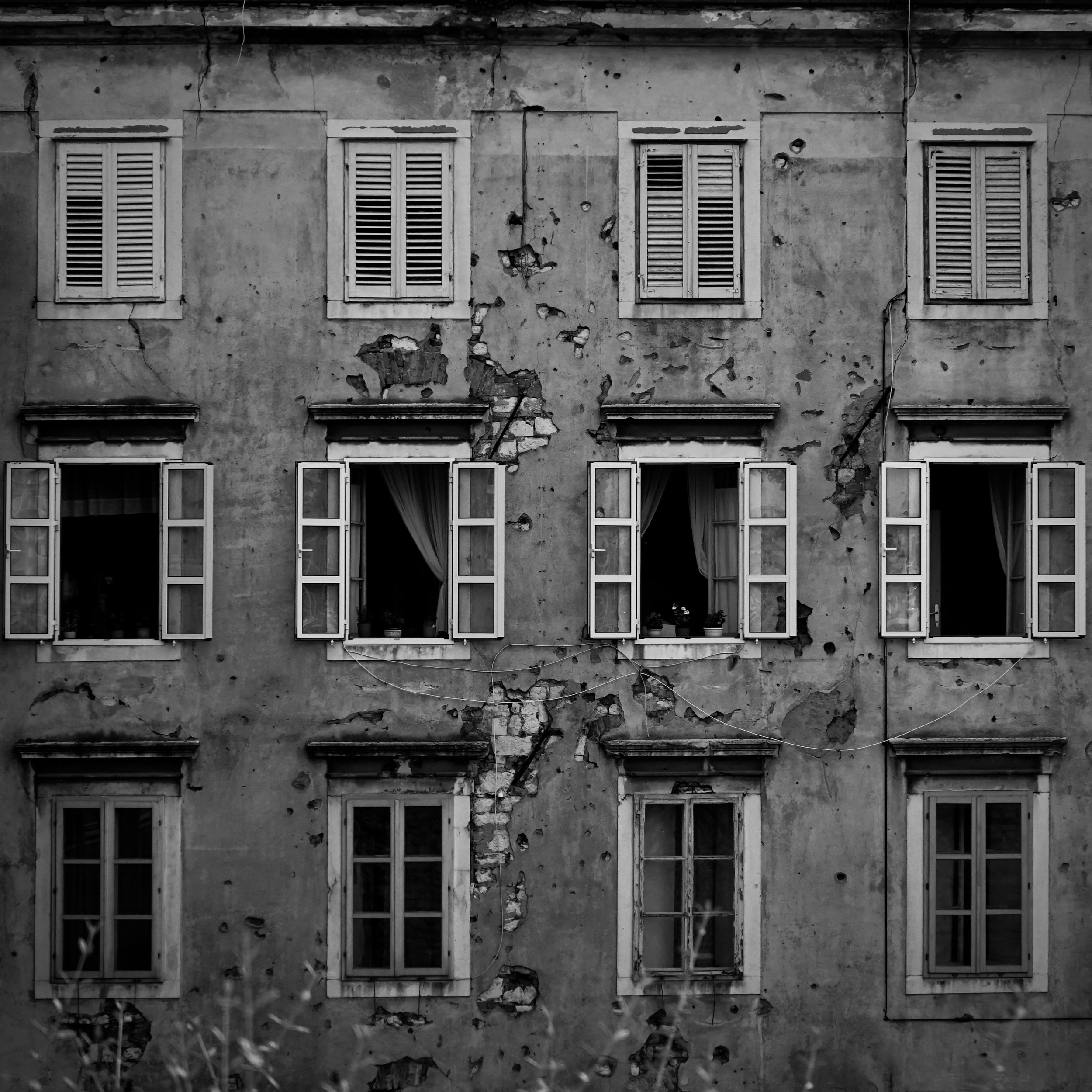 Black and white photo of an old, weathered building with nine windows, some open with curtains, and visible cracks and damage to the wall.