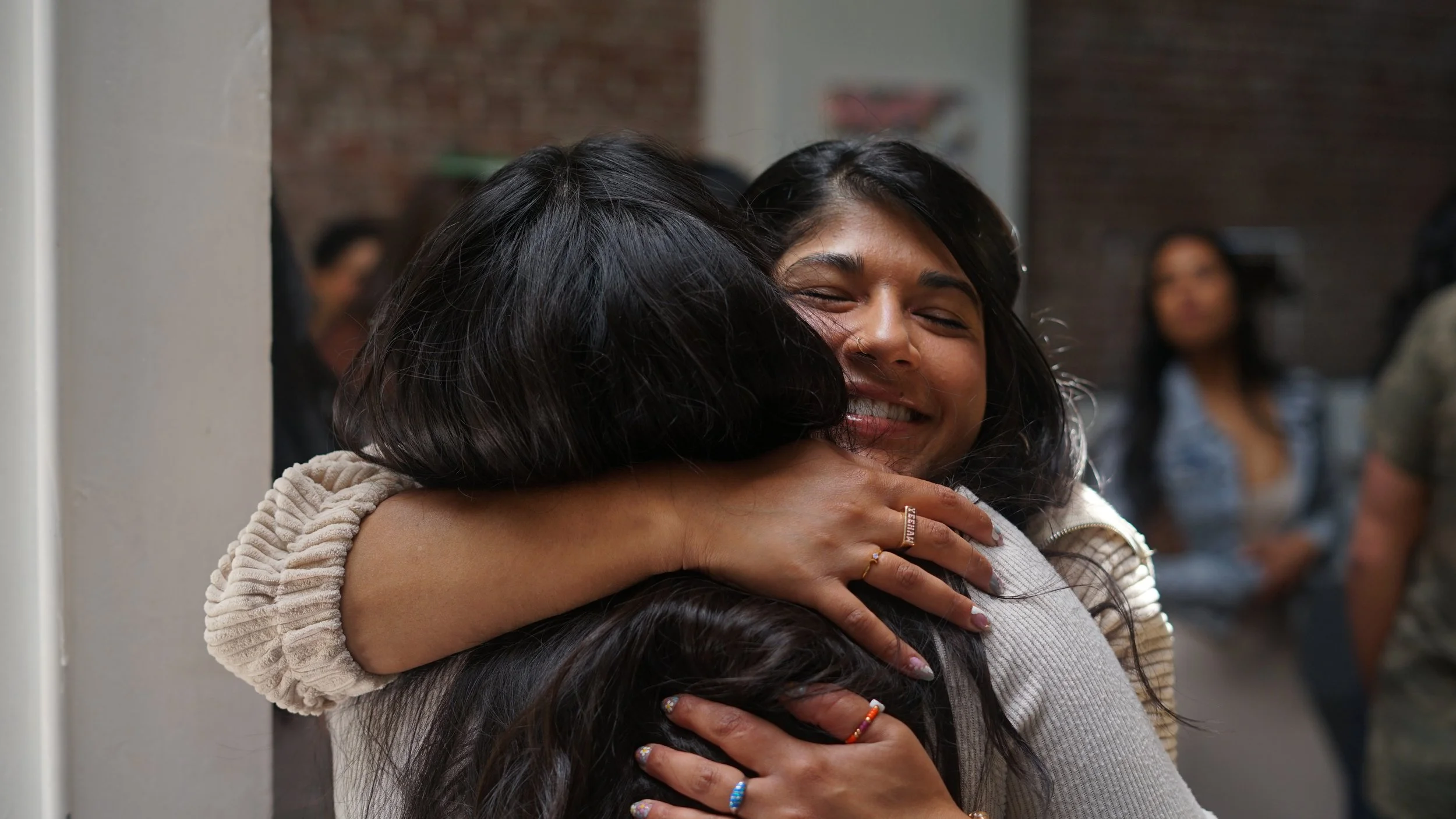 Two women hugging each other, smiling with eyes closed. One woman wears a beige sweater, and the other has long dark hair and is wearing multiple rings.
