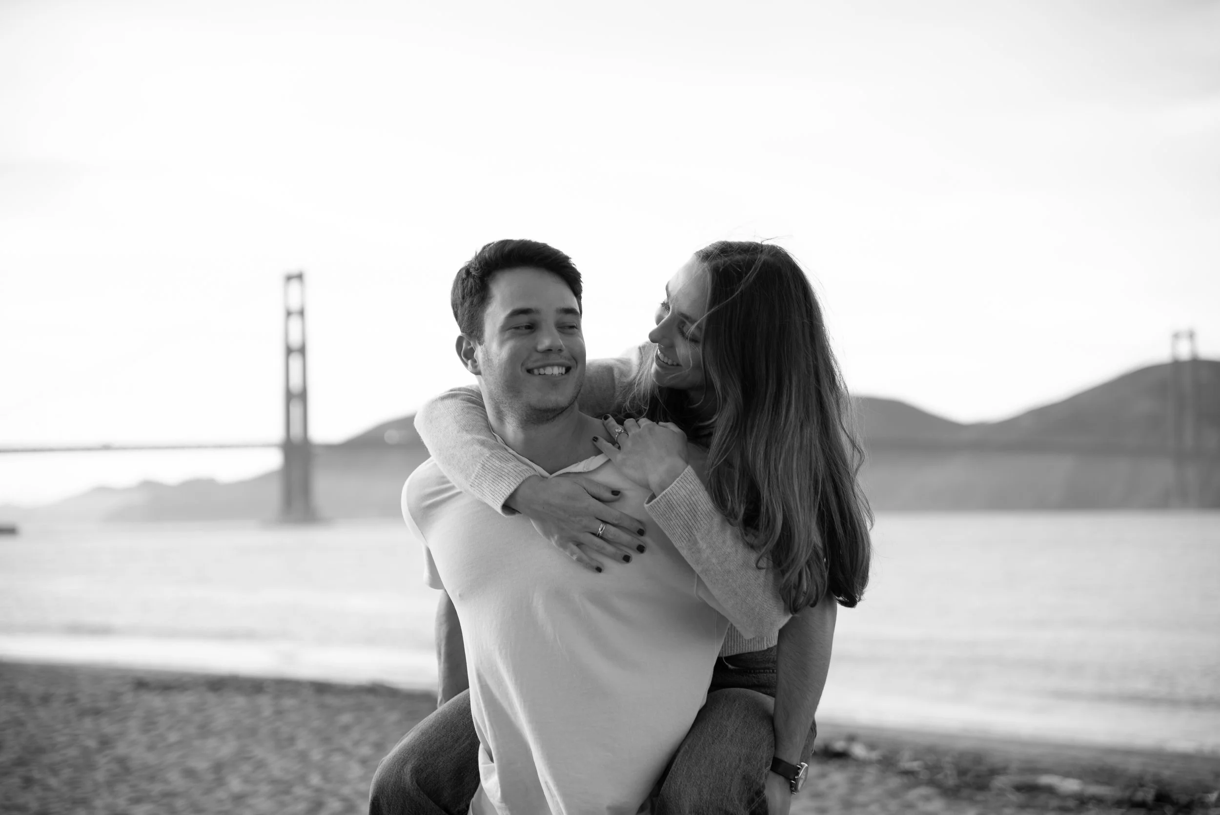 A young man and woman are smiling and embracing outdoors with a bridge in the background.