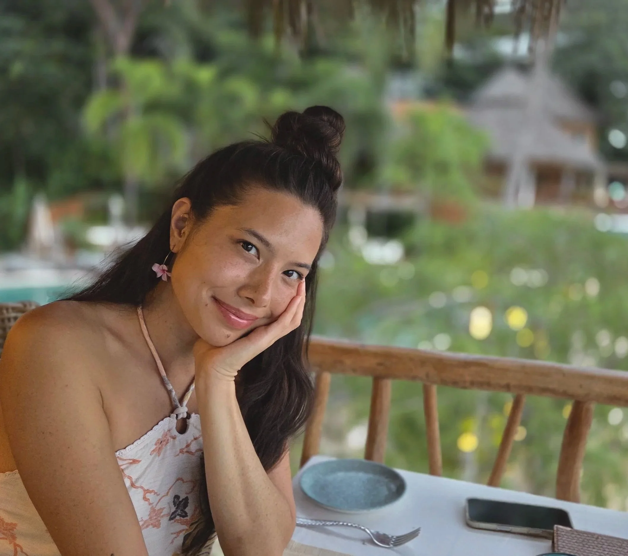 A woman with long dark hair styled in a half-up bun, smiling gently with her head resting on her hand, sitting at a table outdoors with a blurred green natural background.