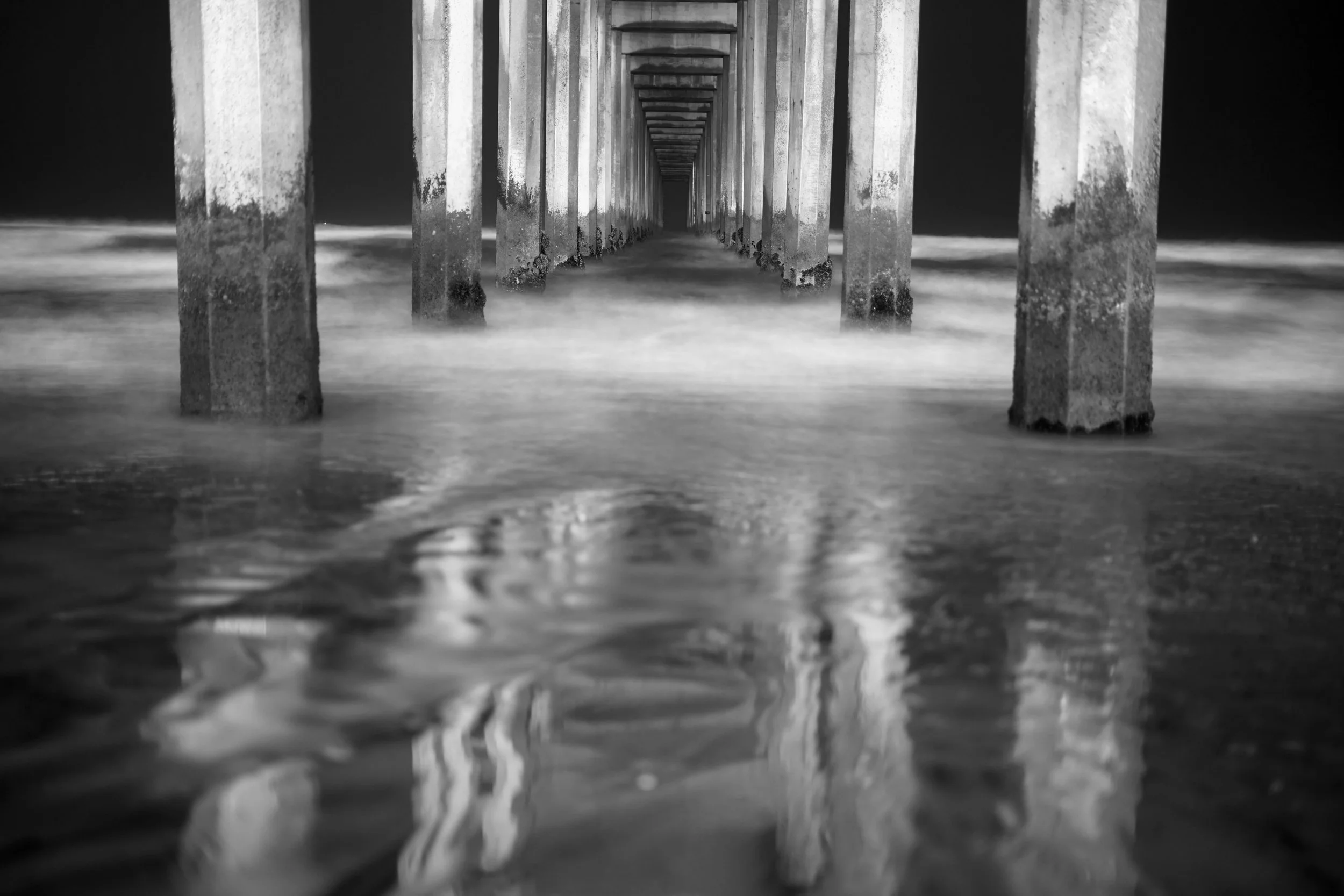 Black and white photo of a pier extending into the ocean with waves crashing against the supports.