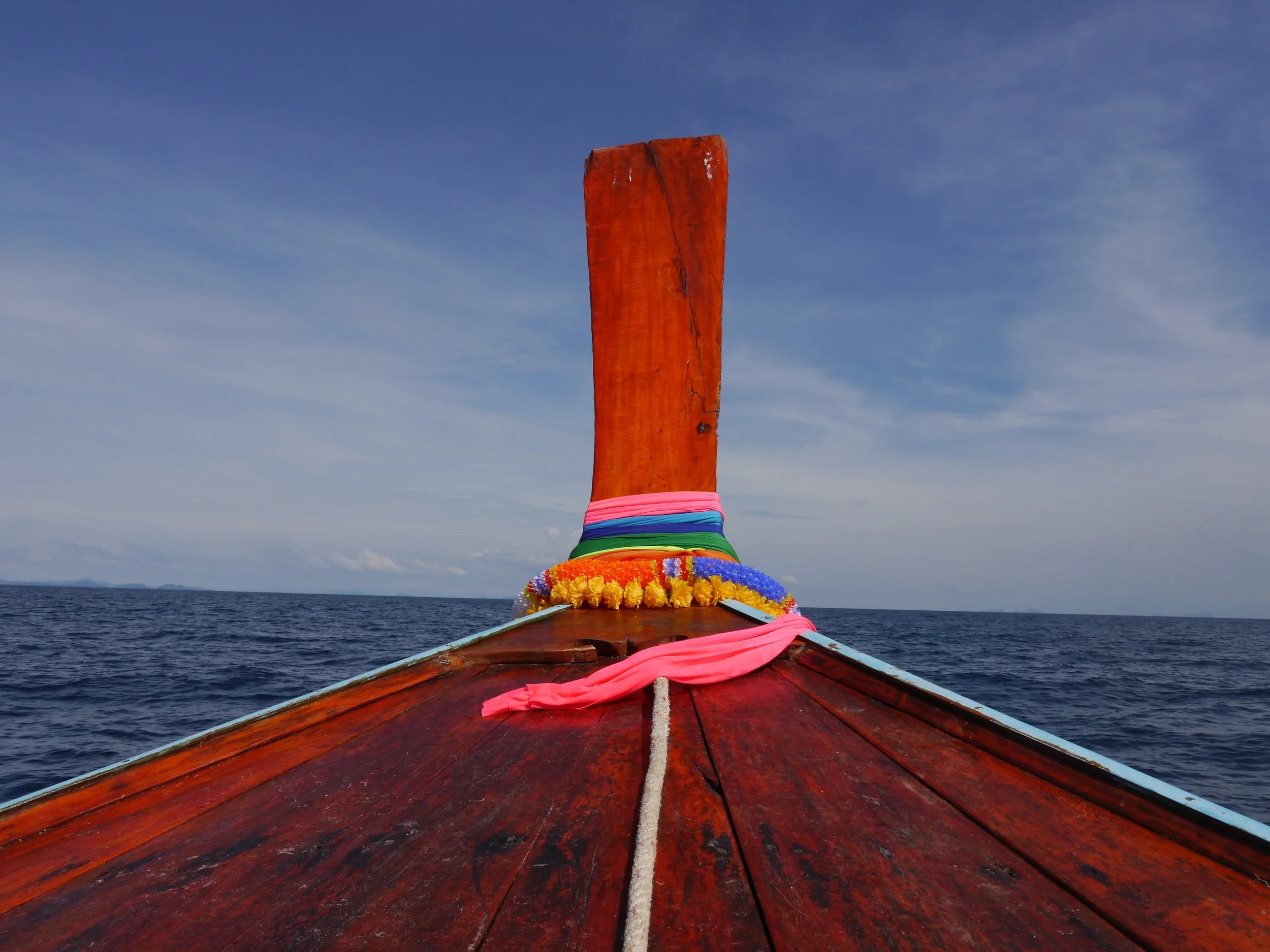 View from the front of a traditional wooden boat on the open sea, with a clear sky and distant land on the horizon, decorated with colorful cloth and flowers at the bow.
