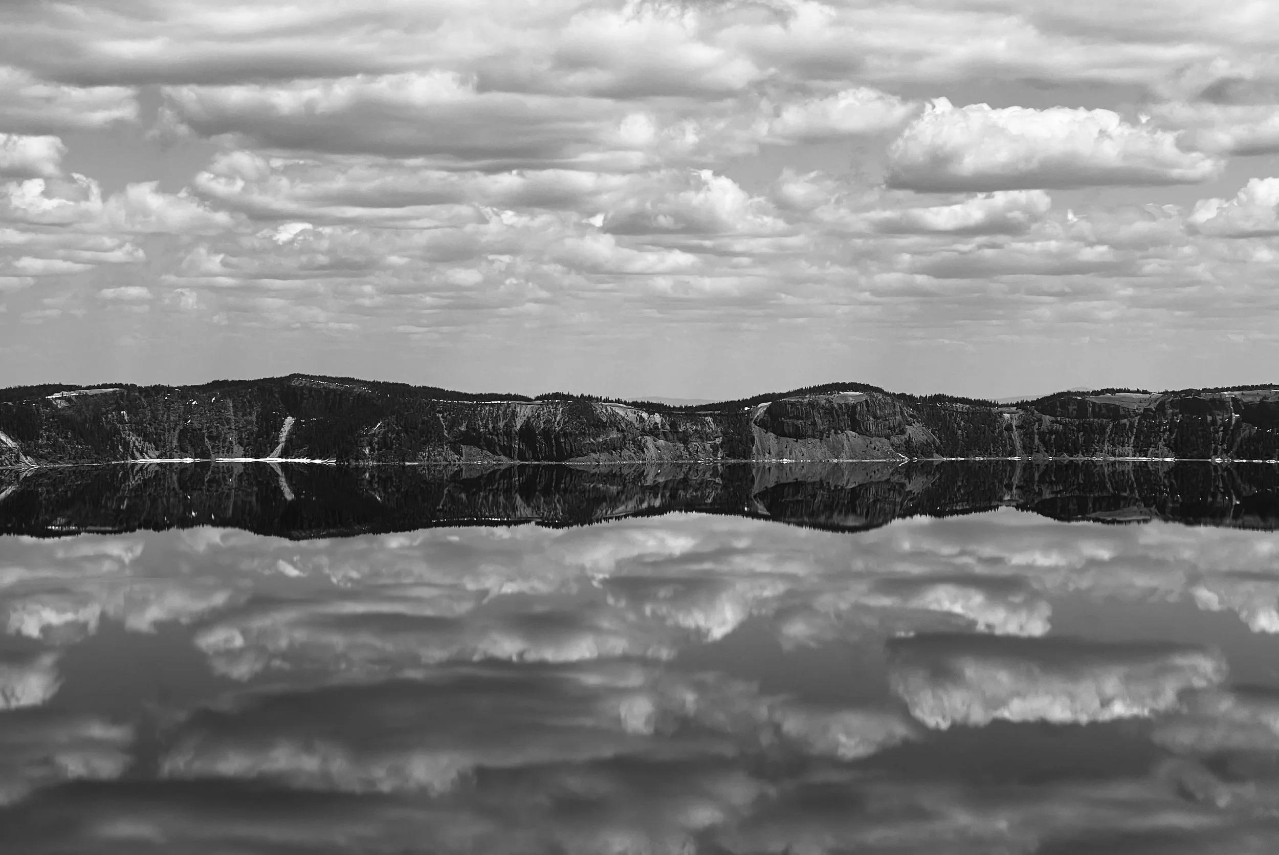 Black and white landscape photo of a lake with calm water reflecting a cloudy sky and distant hills.