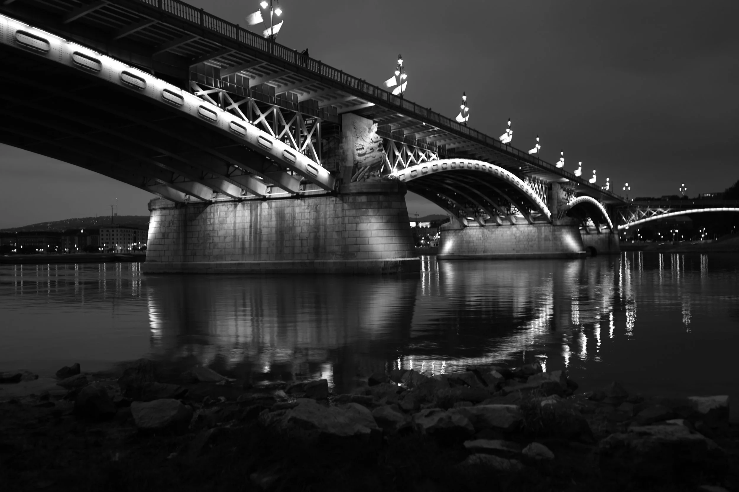 Black and white photo of a large bridge over a body of water, with city lights and buildings in the background, at night.