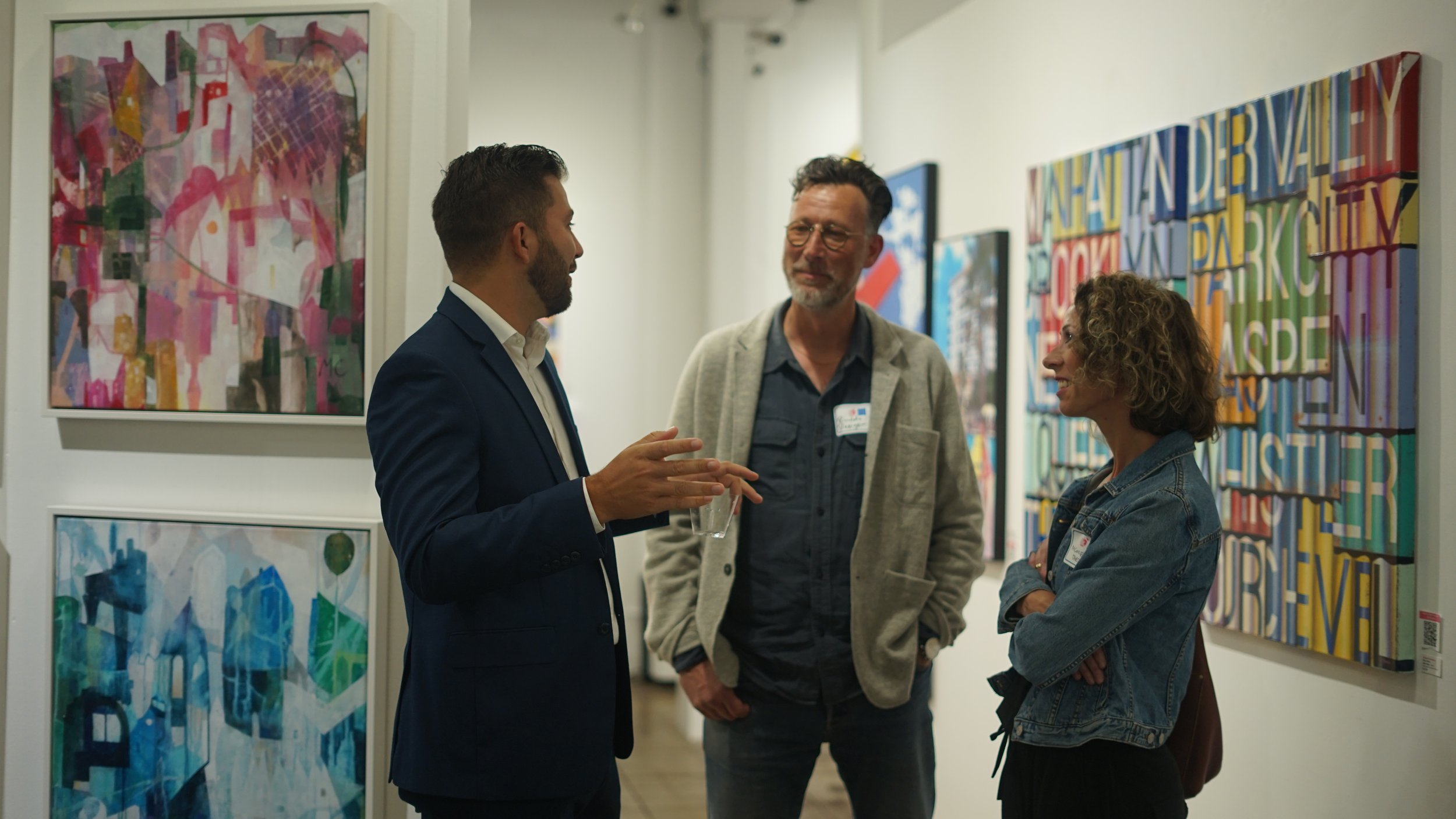 Three people having a conversation in an art gallery, surrounded by abstract paintings on the walls.