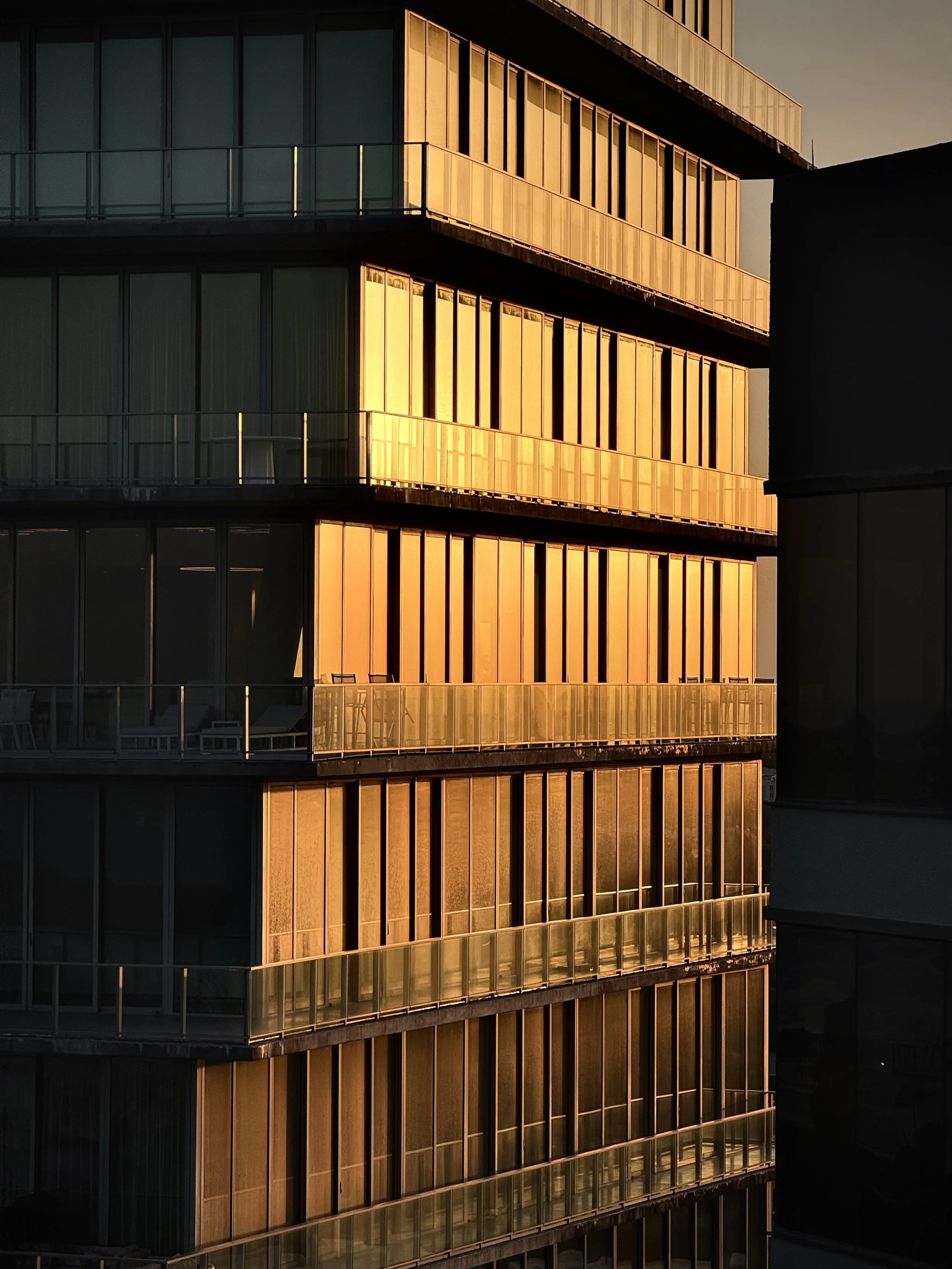 Modern multi-story building with glass windows reflecting the sunset, featuring balconies and vertical architectural details.