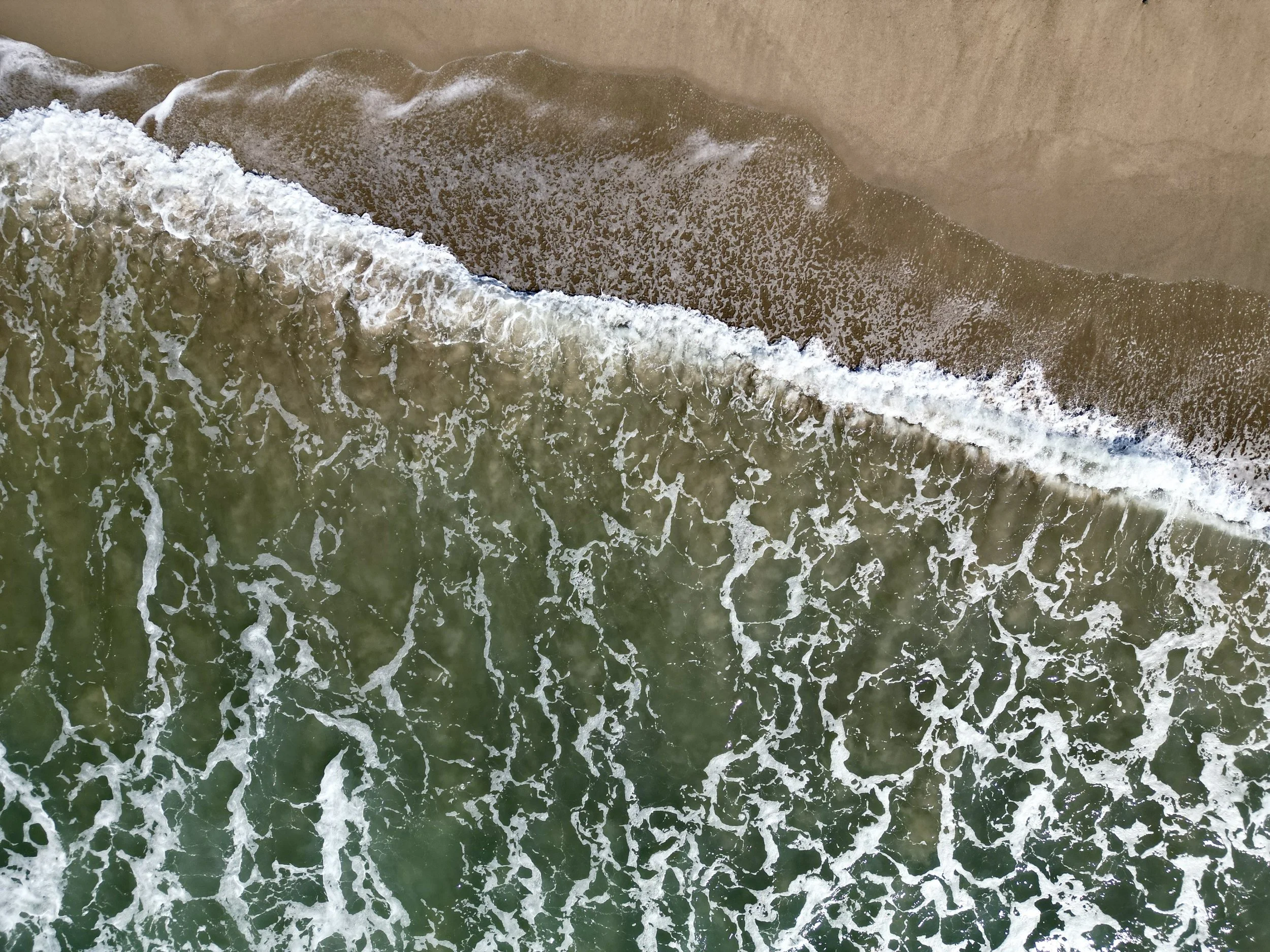 An aerial view of a shoreline with waves crashing onto the beach, showing greenish water and wet sand.
