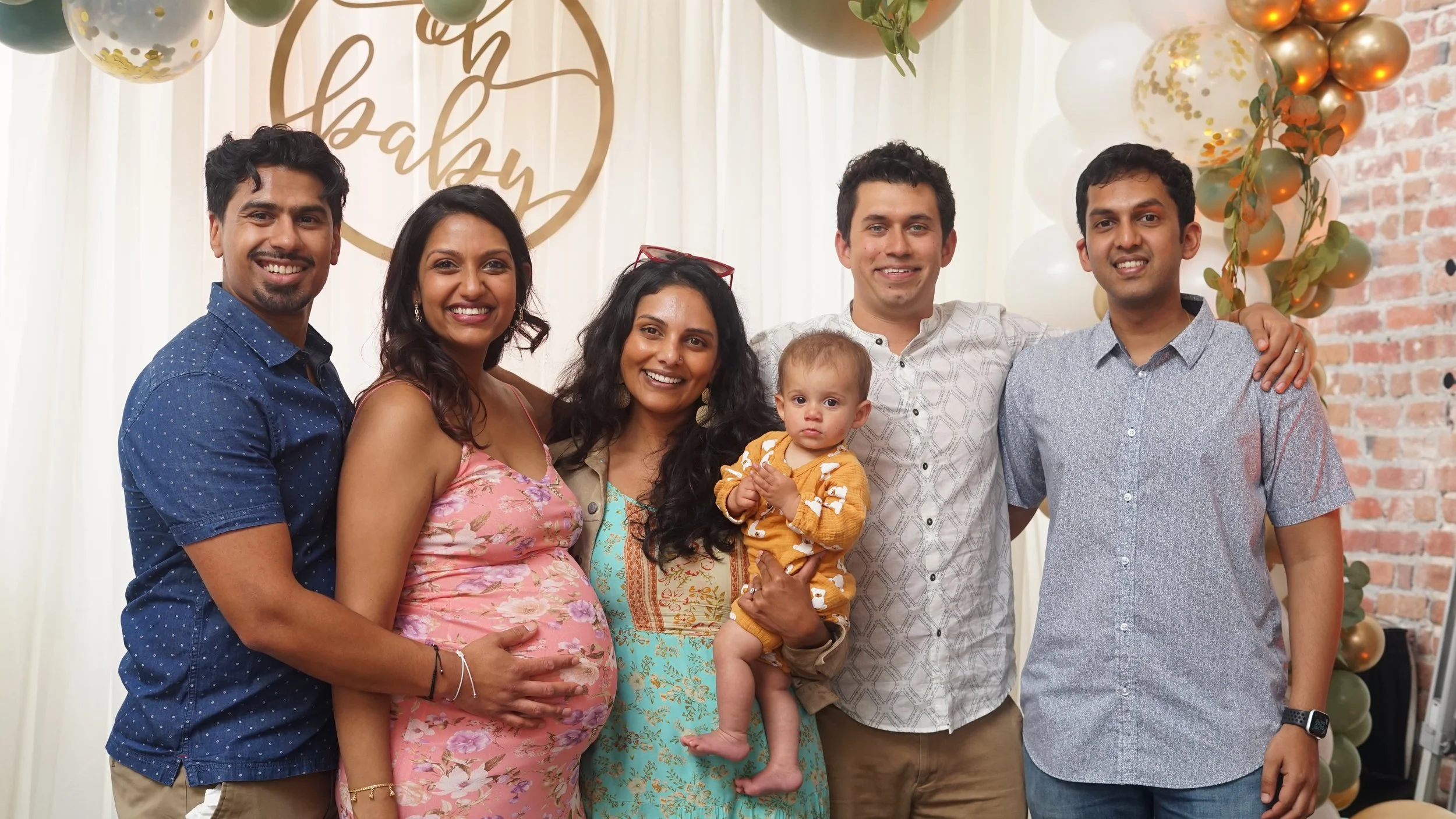 Group of six adults and one baby posing together at a celebration with a decorated balloon backdrop and a sign that says 'oh baby' in the background.