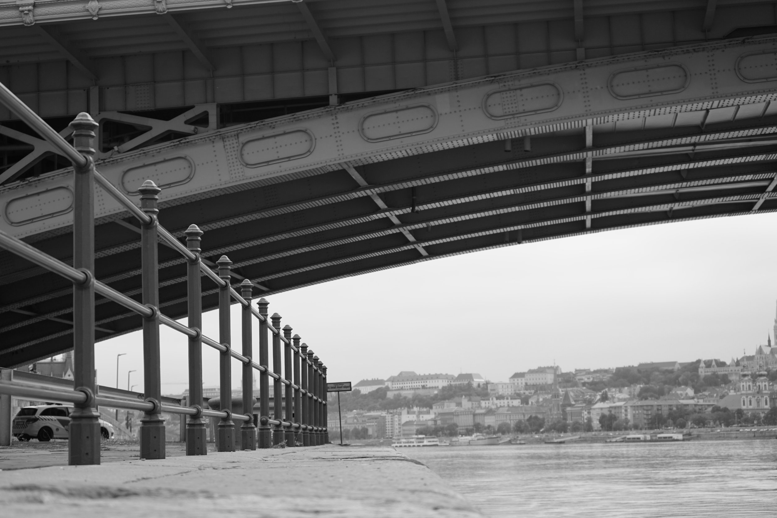 Black and white photograph of an overpass bridge over a river with a cityscape in the background and a fenced walkway in the foreground.