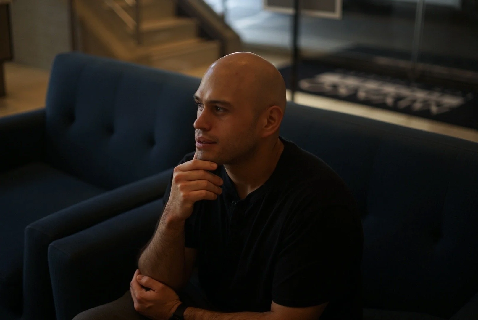 A bald man sitting on a blue couch in a dimly lit room, resting his chin on his hand and looking thoughtfully to the side.