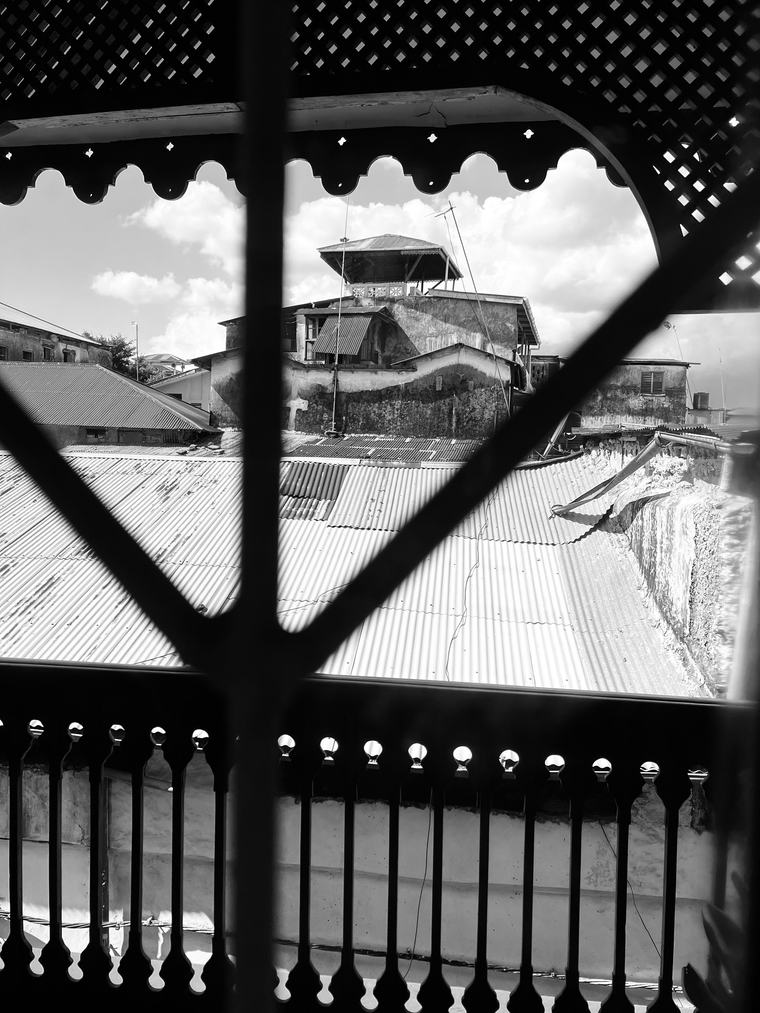 View of rooftops with corrugated metal sheets through a balcony railing, showing an old, weathered building with a small balcony or platform on top, under partly cloudy skies.