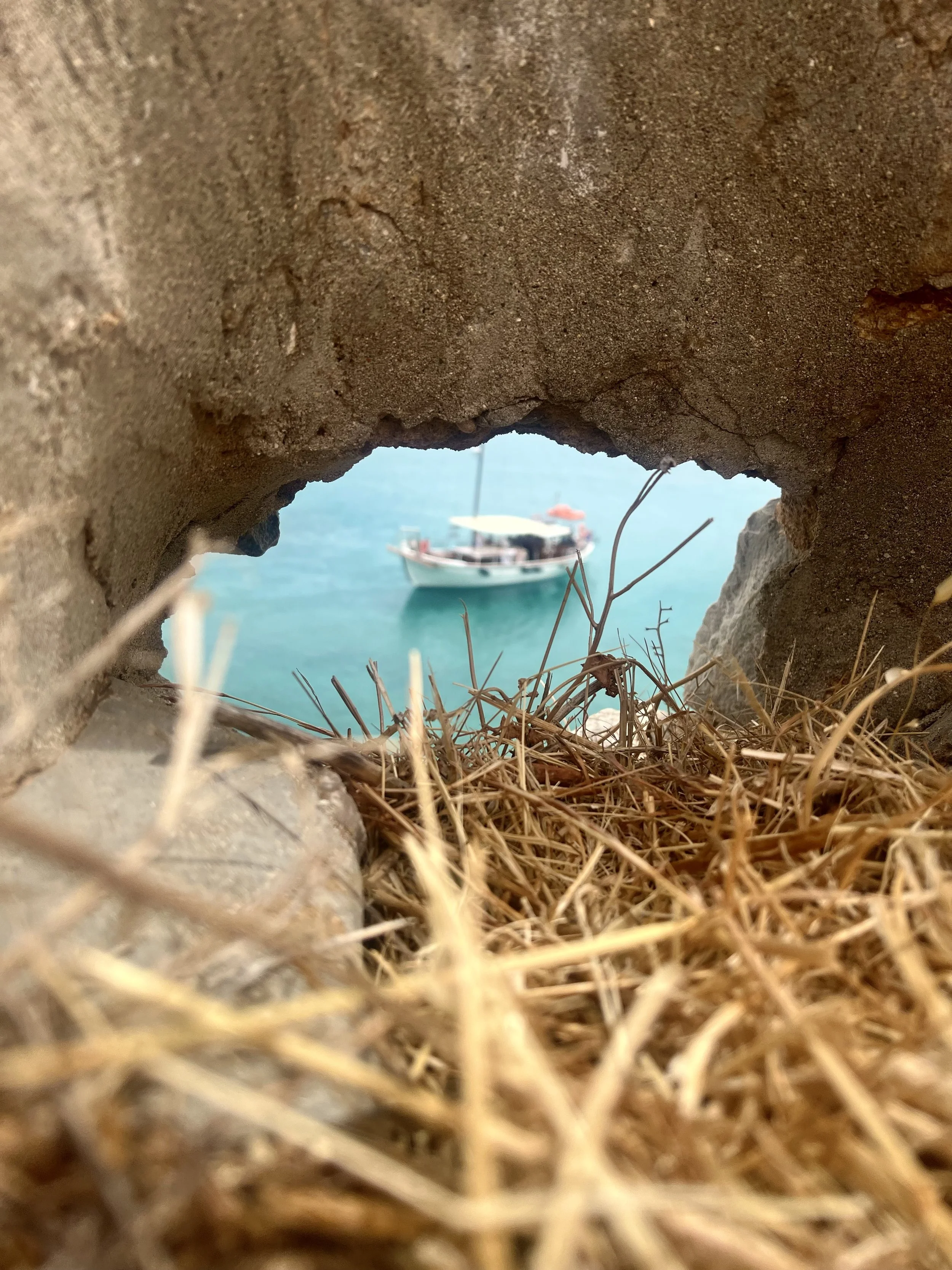View of a boat on the water seen through a small hole in a brick or stone structure, with dry grass in the foreground.