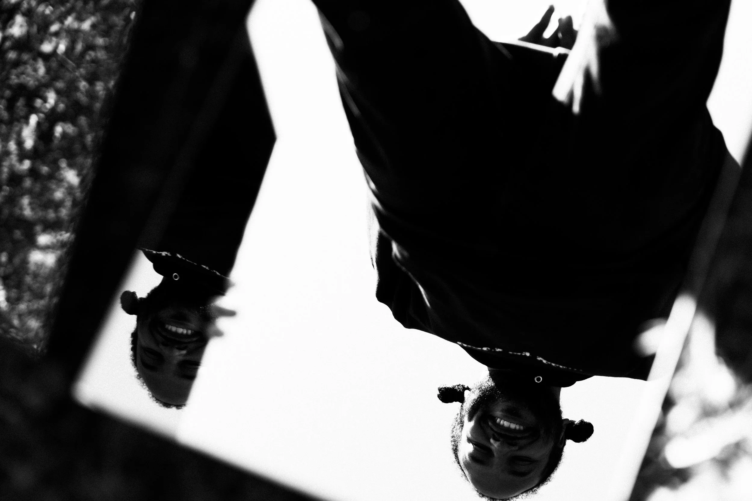 A black and white photo of a young woman looking at her reflection in a mirror, smiling and wearing a baseball cap. She has curly hair and is outdoors with trees in the background.