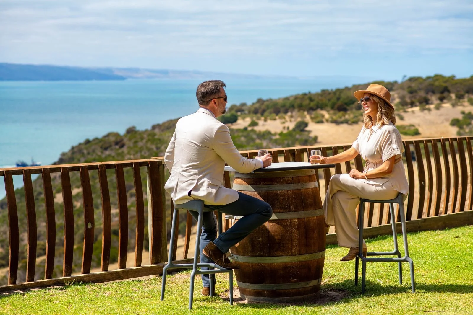 A man and a woman sitting at a round table made from a barrel, enjoying drinks outdoors with a scenic view of a lake and hilly landscape in the background, during daytime.