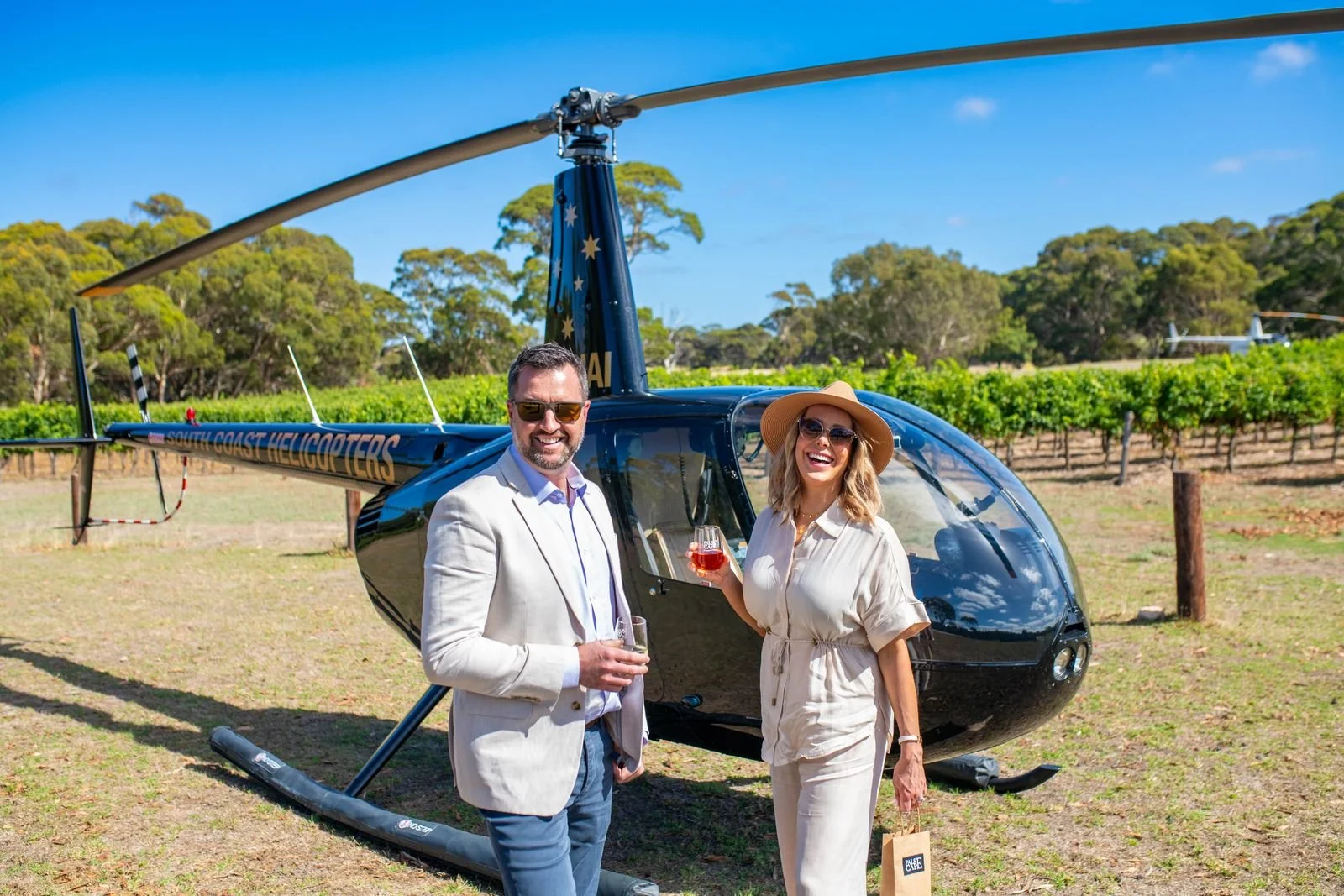 A man and a woman stand in front of a black helicopter at a vineyard, holding glasses of wine and smiling.