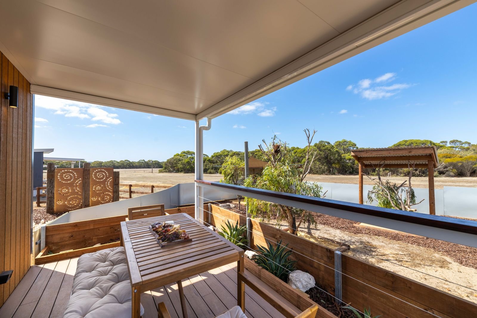 Cozy outdoor balcony with wooden furniture, plants, and scenic landscape of trees and open field under blue sky.