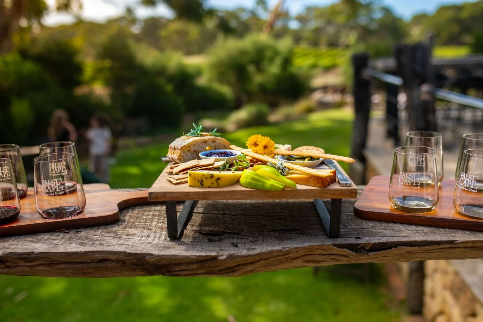 A wooden table with a serving tray of cheese, crackers, sliced fruit, and small bowls of jam and pickles outdoors with a blurred background of greenery and two people in conversation.